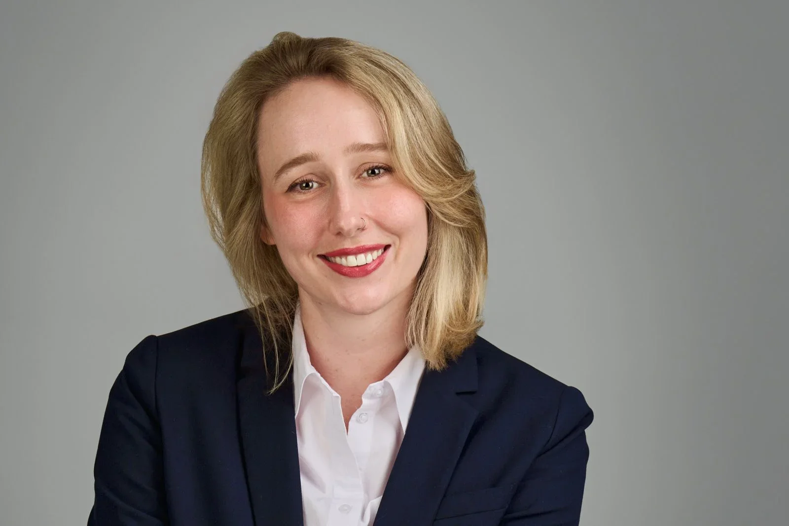 A woman with shoulder-length blonde hair, wearing a navy blazer over a white shirt, smiling against a plain gray background.