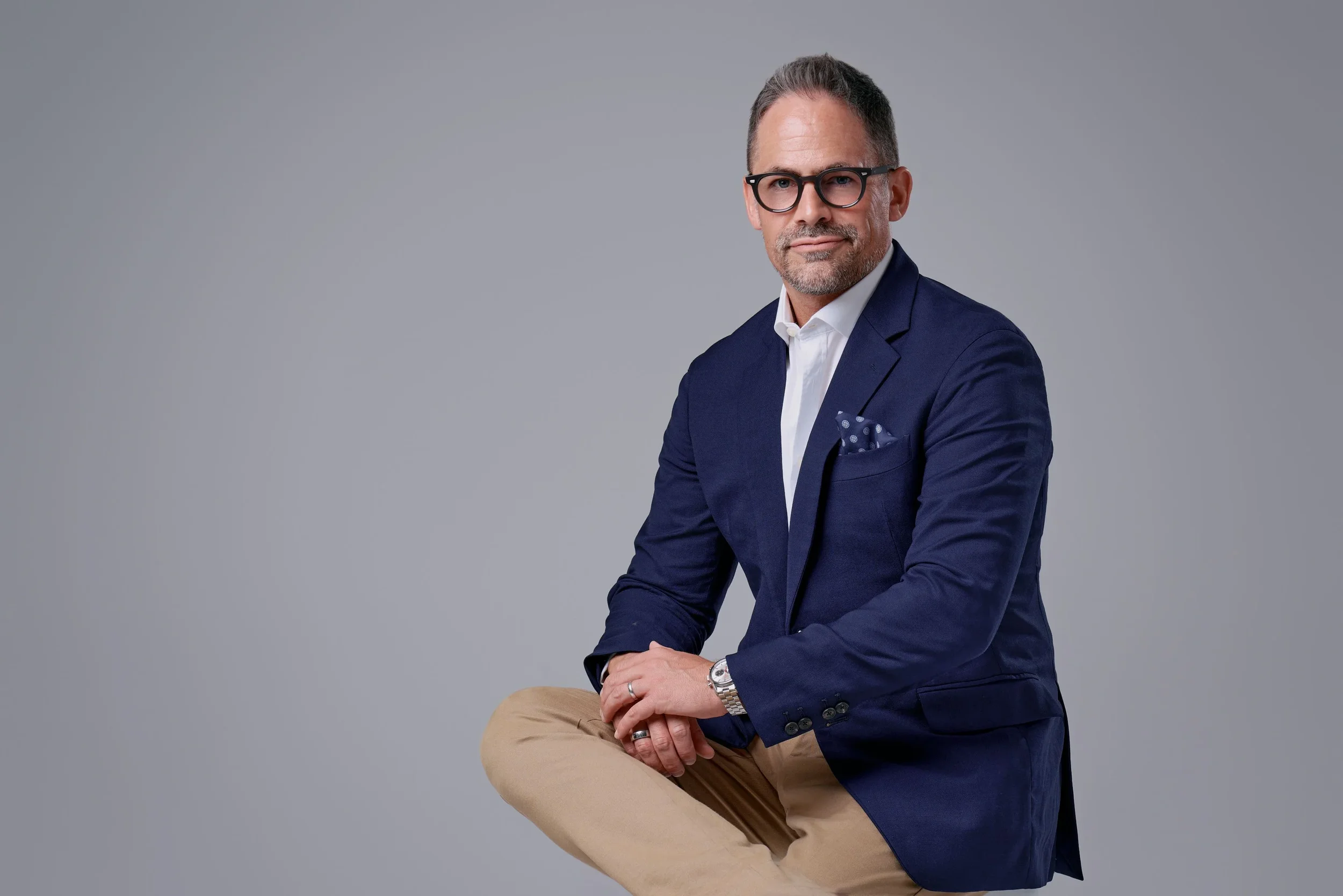 A young man with glasses wearing a dark blazer over a white shirt, red pants, and black shoes, sitting on a white stool against a plain gray background, smiling at the camera.