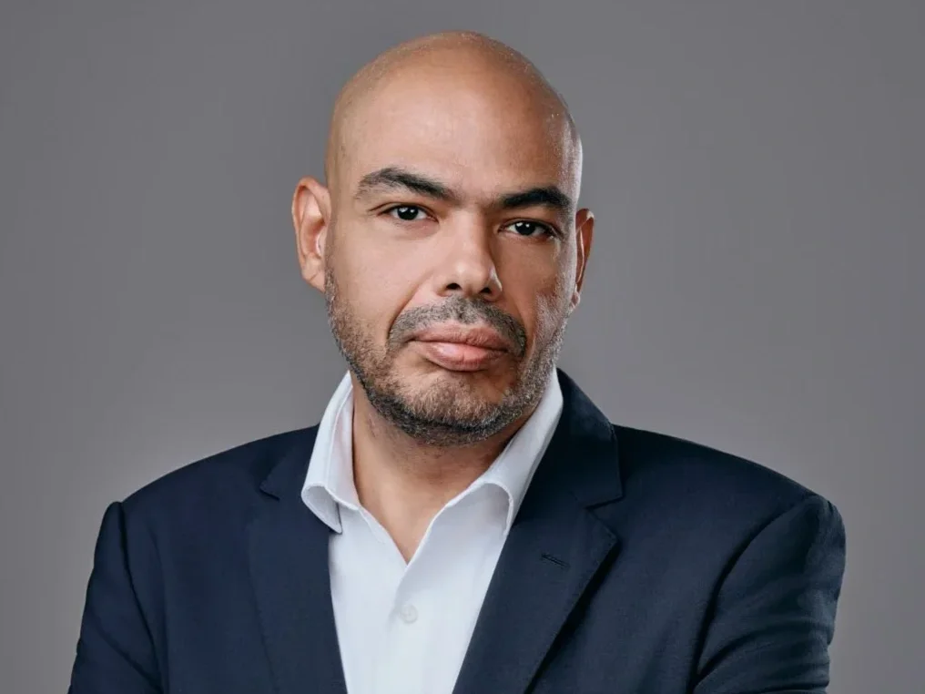 A middle-aged man with a beard and shaved head, dressed in a navy blazer and white shirt, posing against a plain gray background.