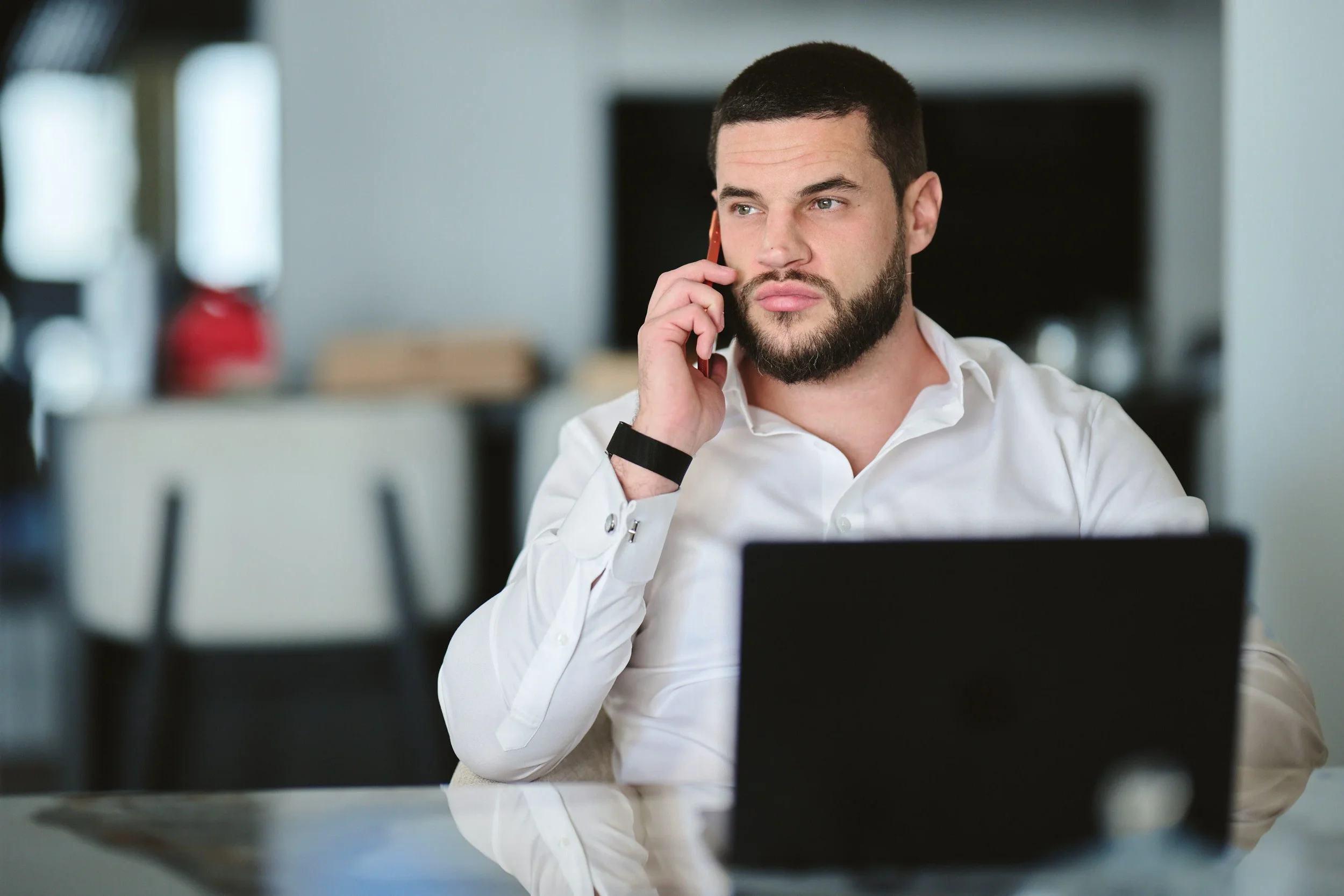A man with dark hair and a beard, dressed in a white shirt, is sitting at a desk in an office, talking on a cell phone.