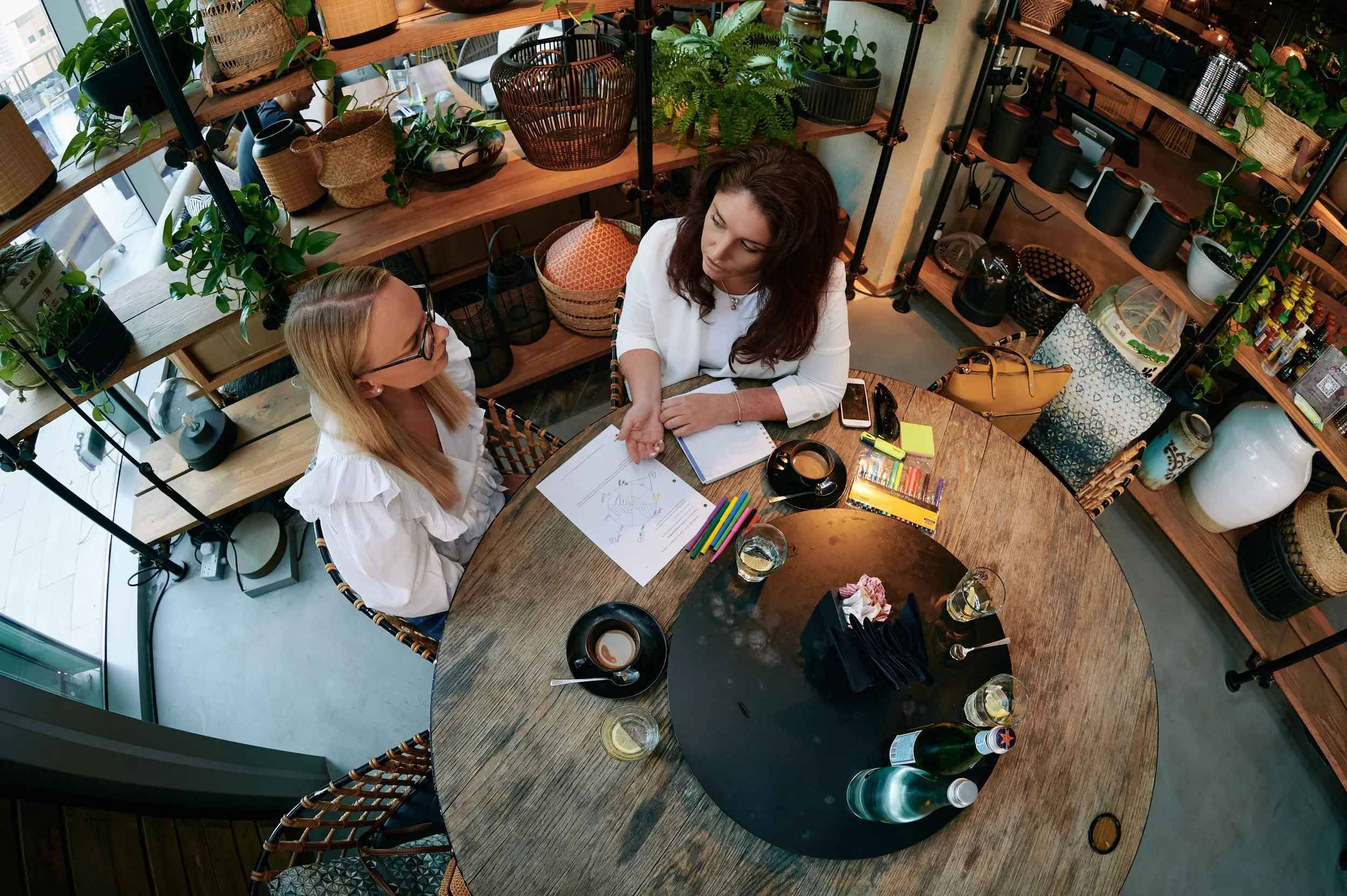 Two women seated at a round wooden table in a cozy, plant-filled café or restaurant, engaged in conversation with notebooks, colorful pens, and drinks on the table.