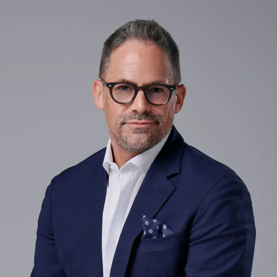 A middle-aged man with gray hair and glasses wearing a navy blue suit and white shirt posing against a gray background.
