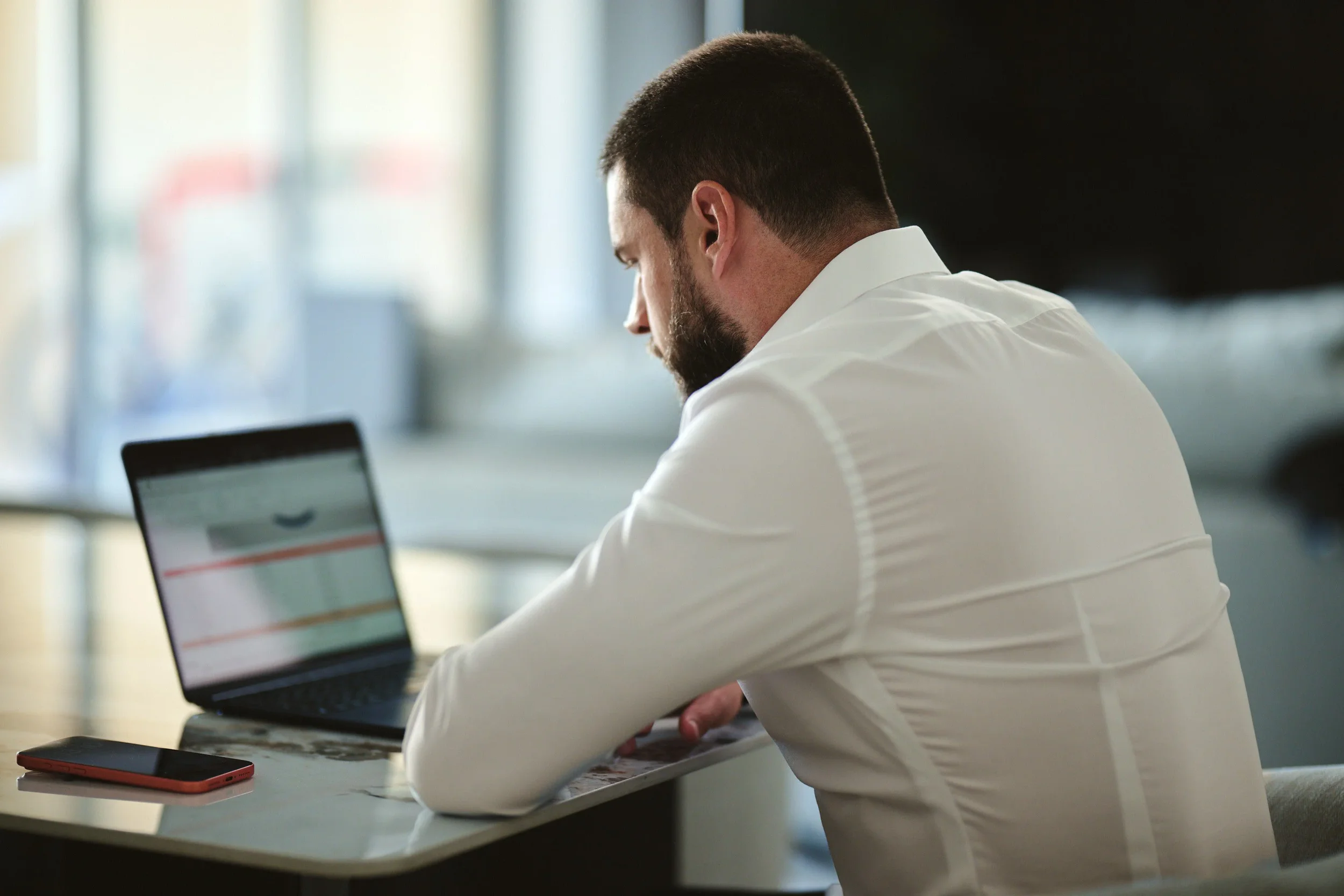 A man with dark hair and a beard, dressed in a white shirt, sitting at a table using a laptop. A smartphone is placed on the table in front of him. The background is blurred with natural light coming through windows.