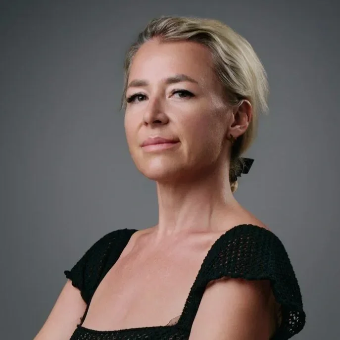 A confident woman with blonde hair tied back, wearing a black, short-sleeved top, standing against a gray background.