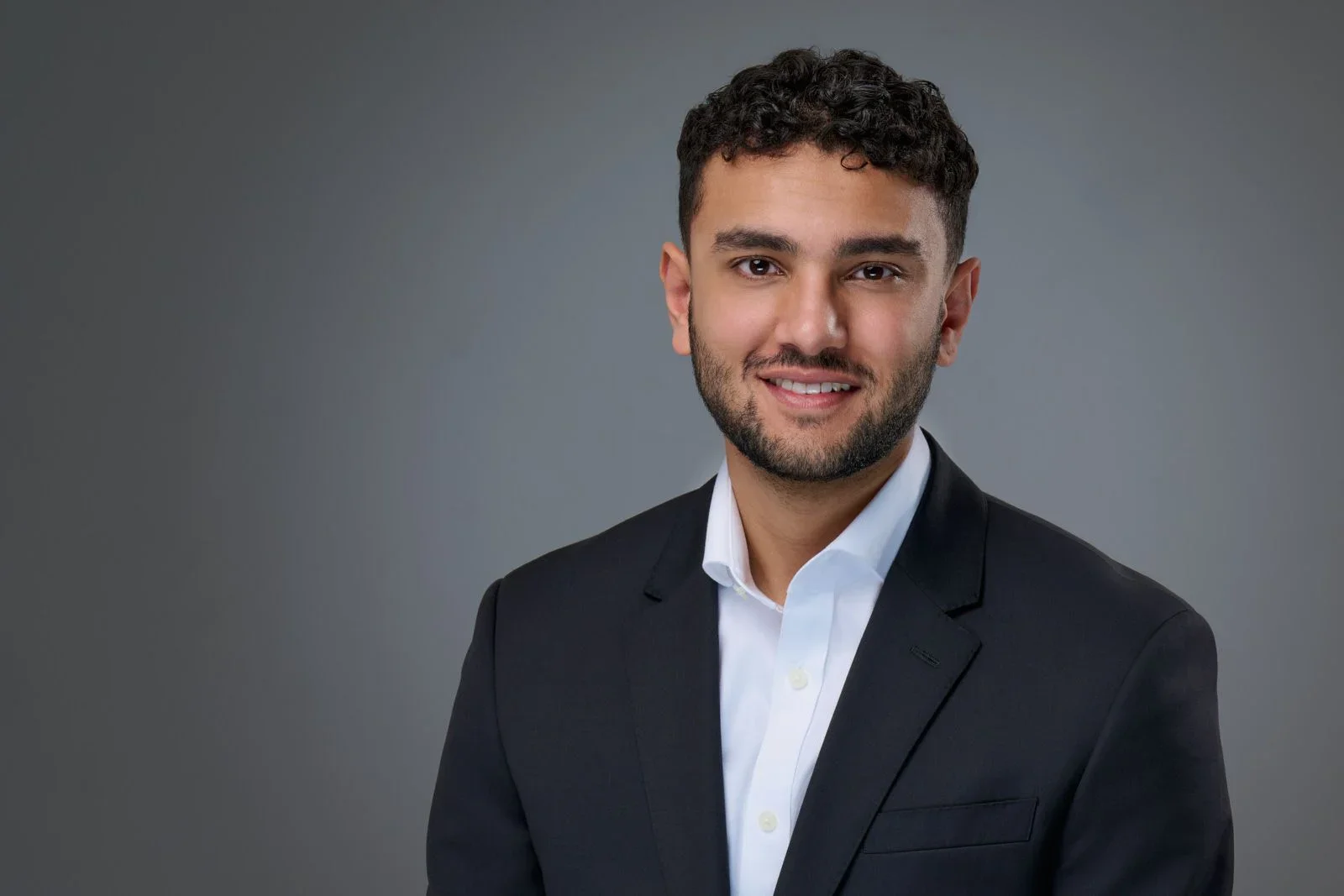 A professional young man with dark curly hair, a beard, and a friendly smile, wearing a black suit jacket and a white dress shirt, posing against a gray background.