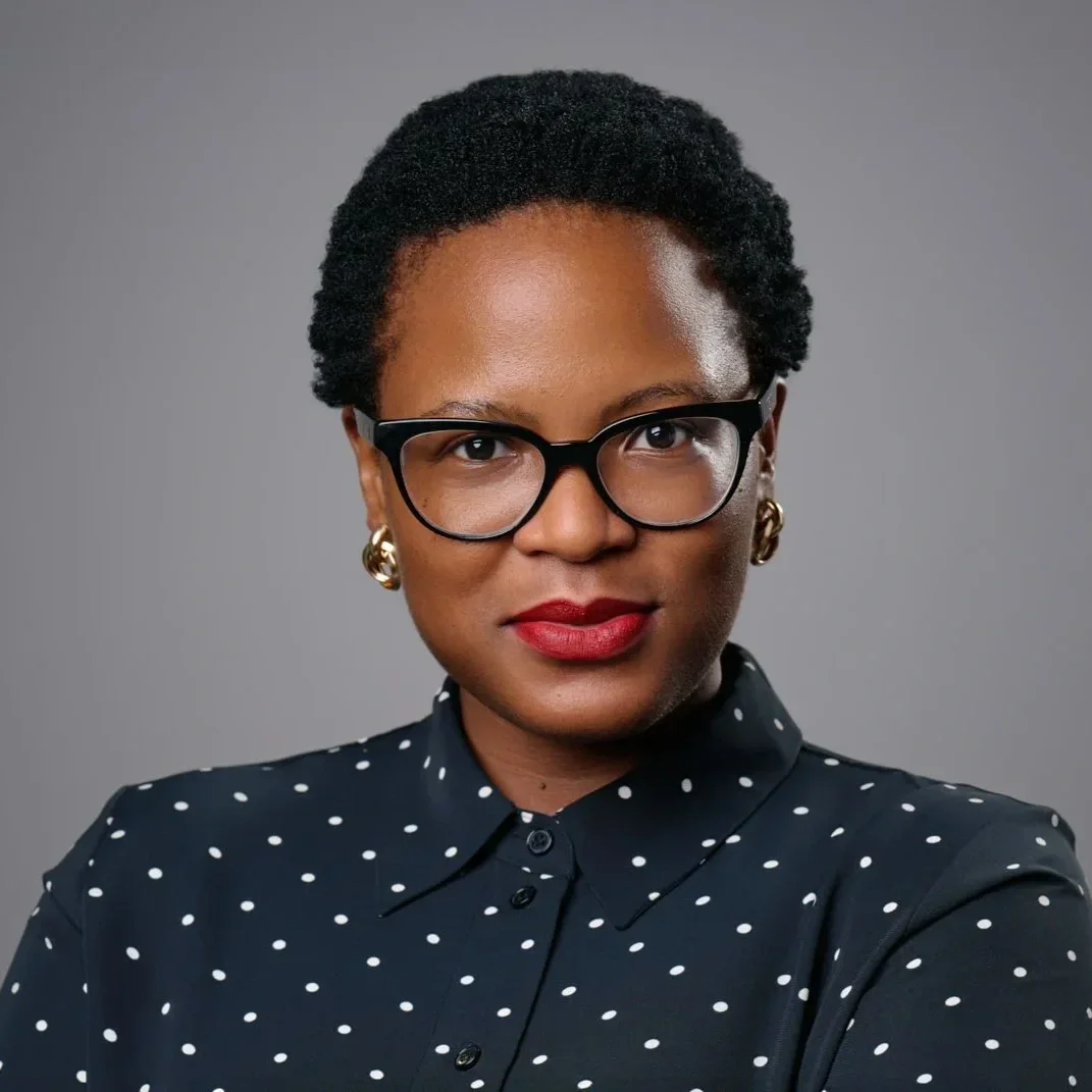 A woman with short curly black hair, wearing black-framed glasses, gold earrings, red lipstick, and a navy blue polka dot blouse, posing against a gray background.