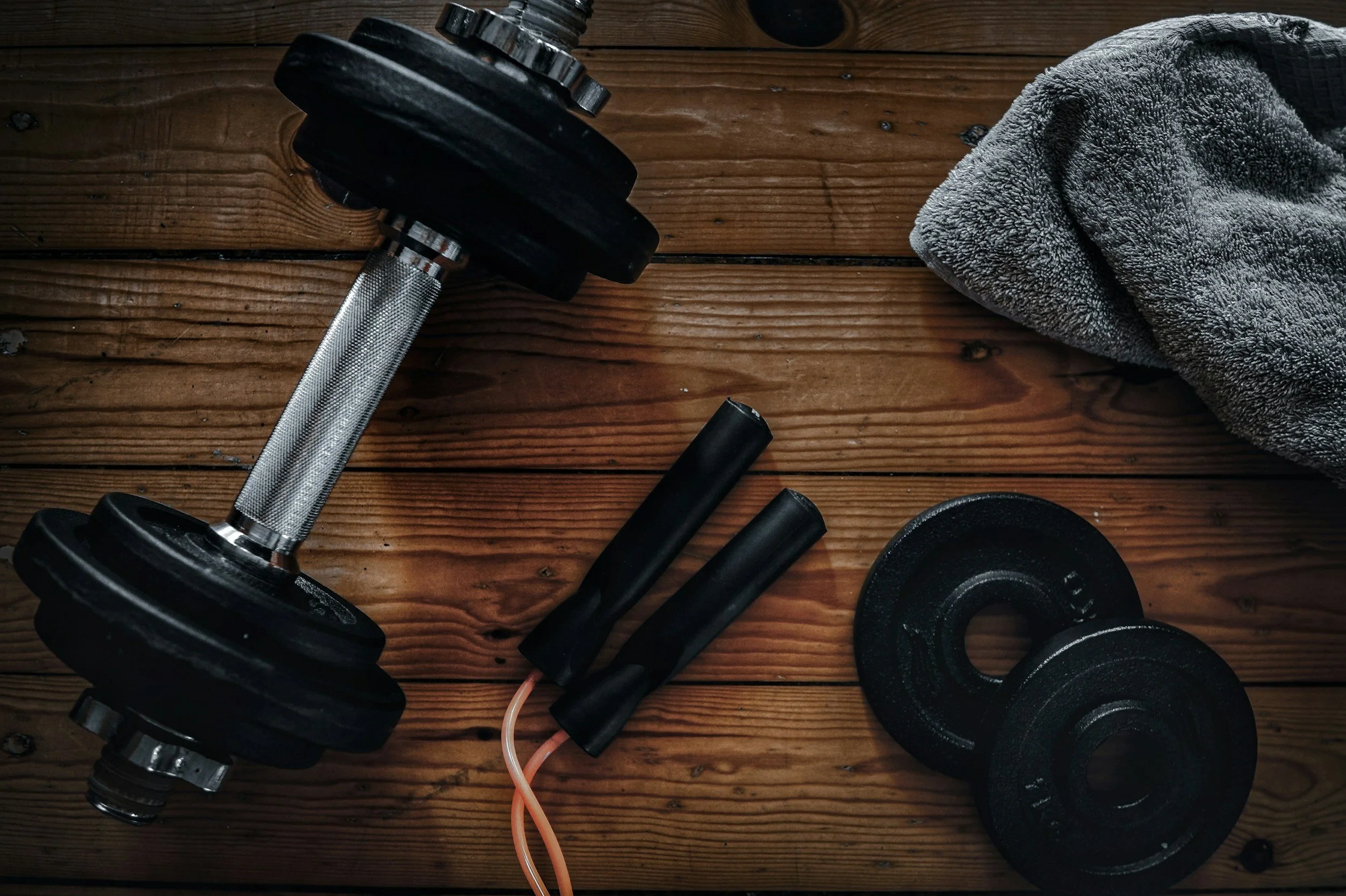 Dumbbell, jump rope, extra weight plates, and a towel on a wooden surface.