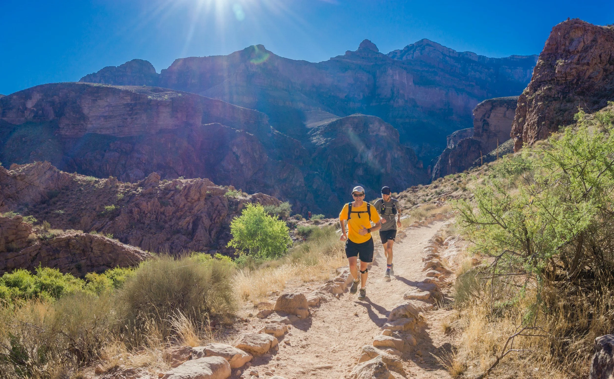 Two hikers, one wearing a yellow shirt and the other in darker clothing, walk along a rocky trail in a canyon with tall cliffs and mountains in the background on a sunny day.