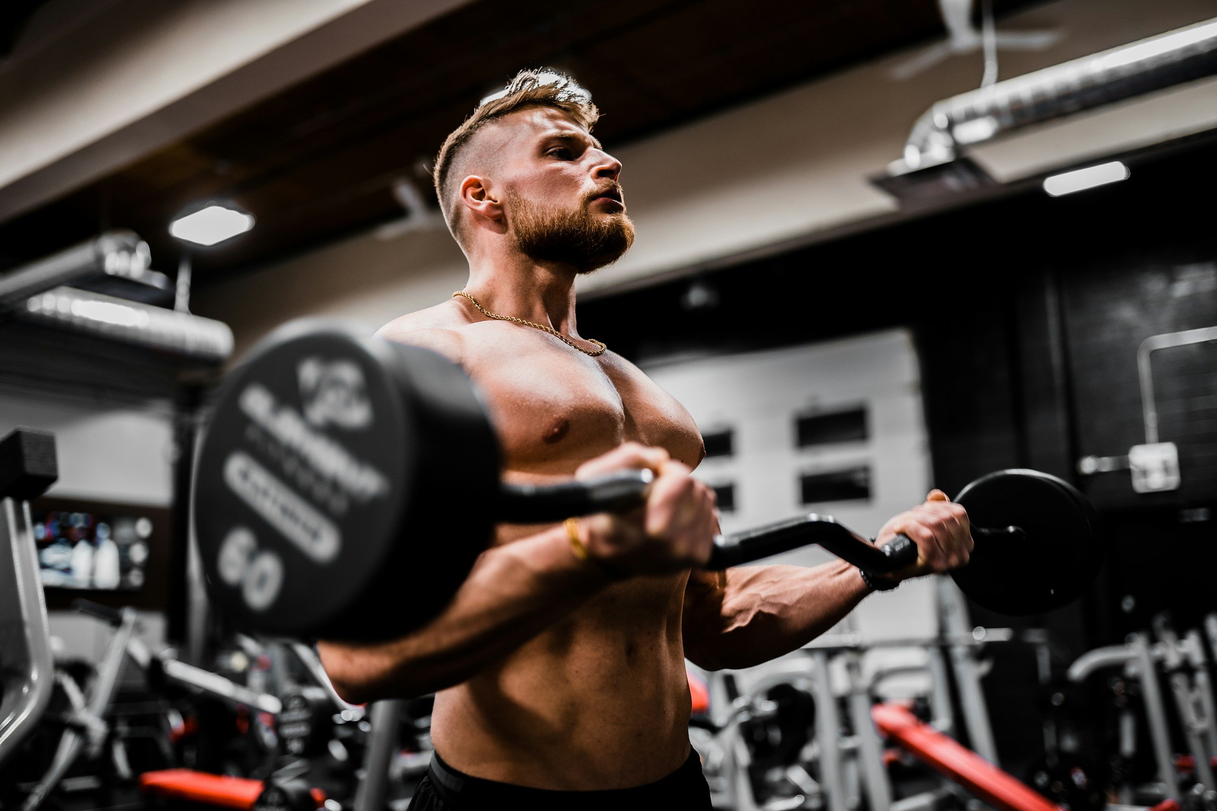A shirtless man with a beard and styled hair lifting weights in a gym.