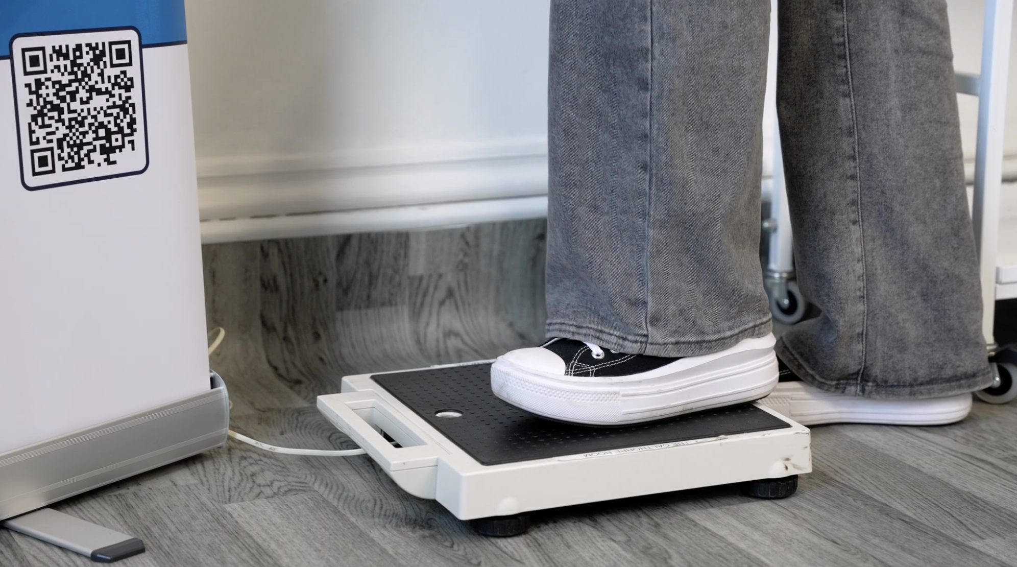 Person standing on a digital weighing scale, with gray pants and black shoes, in a clinical or medical setting.