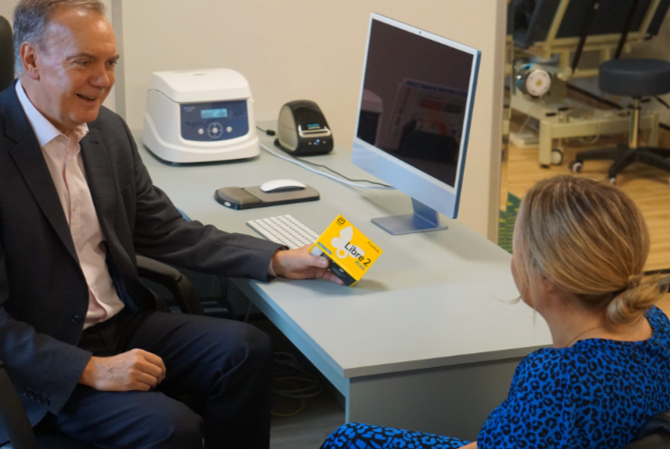 A man in a gray suit hands a woman a yellow box labeled 'Libra' in an office setting with a computer, printer, and other office equipment on the desk.
