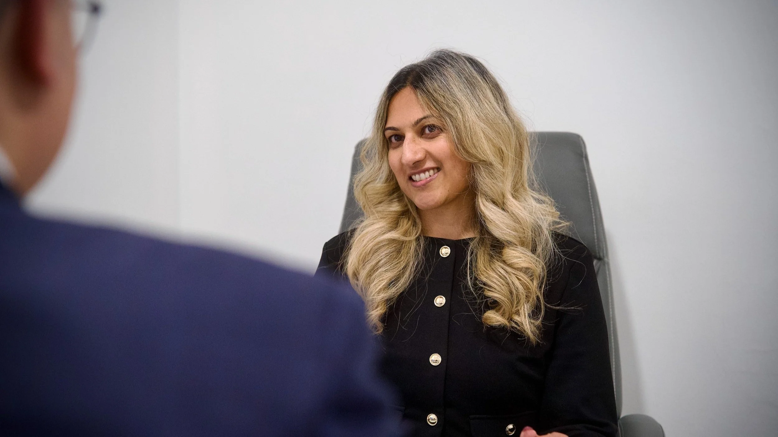 Dr Bavna Kallan smiling while sitting in a chair, engaged in a conversation with a patient whose shoulder is visible in the foreground.