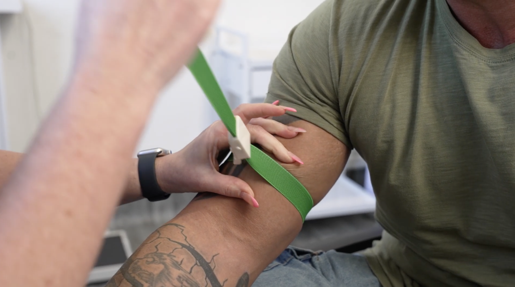A healthcare professional is preparing to draw blood from a patient's arm. A green tourniquet is around the patient's upper arm.