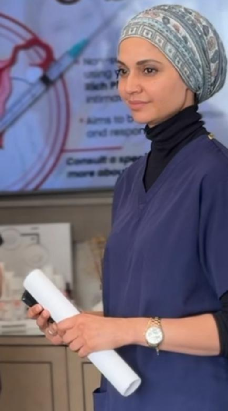 Nurse Nusra Yasmeen in medical scrubs and a patterned headscarf holding a rolled-up document and a marker, standing in front of a medical training presentation screen.