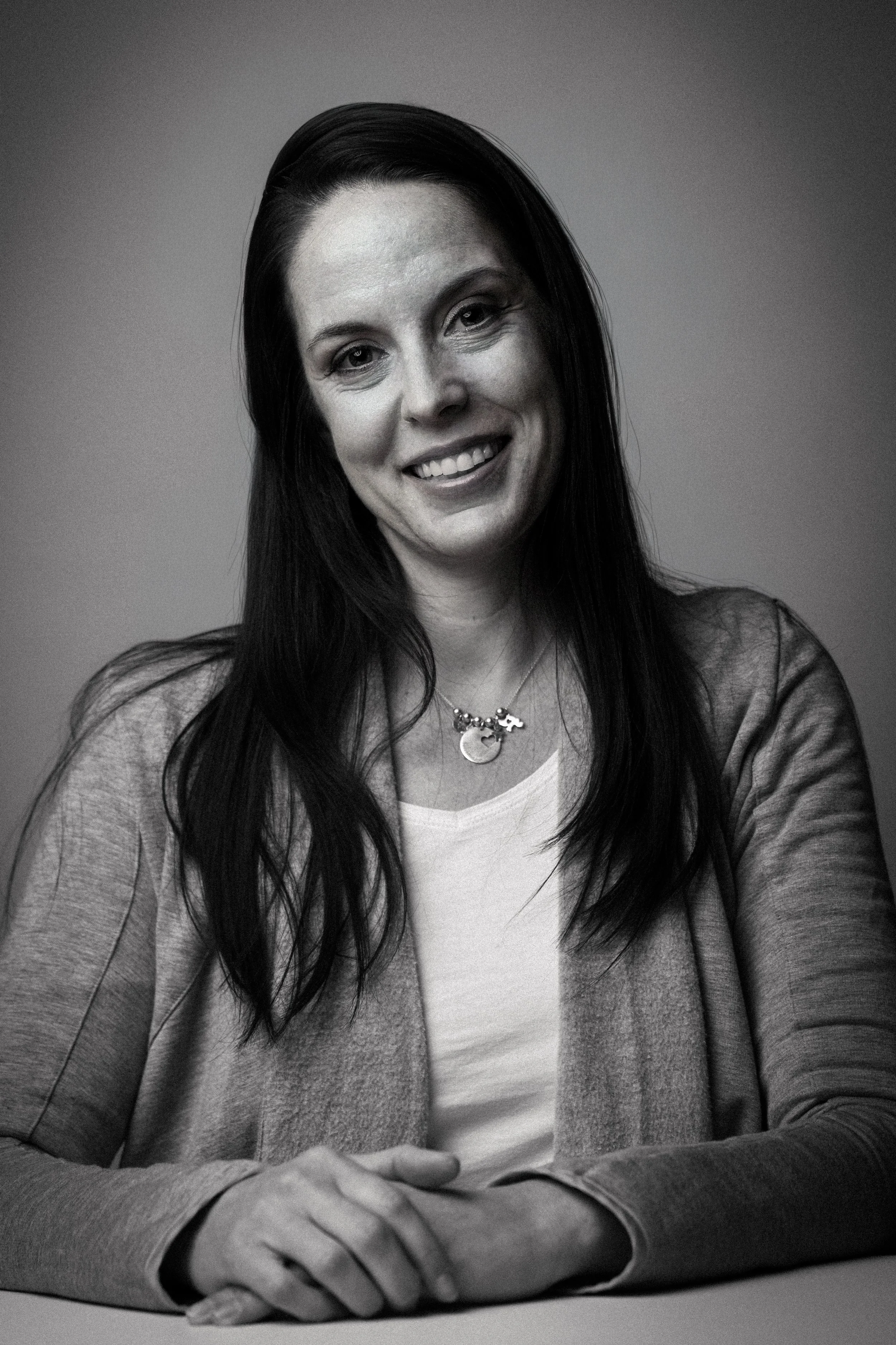 A black and white portrait of a woman at Brand Viva Media with long dark hair, smiling at the camera, wearing a light-colored top, a cardigan, and a necklace with multiple charms.