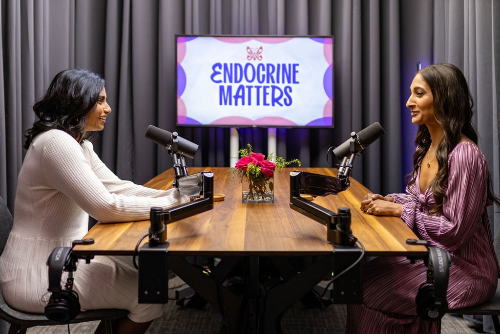 Two women having a podcast discussion in a studio with microphones, a table, and a screen showing 'Endocrine Matters.'