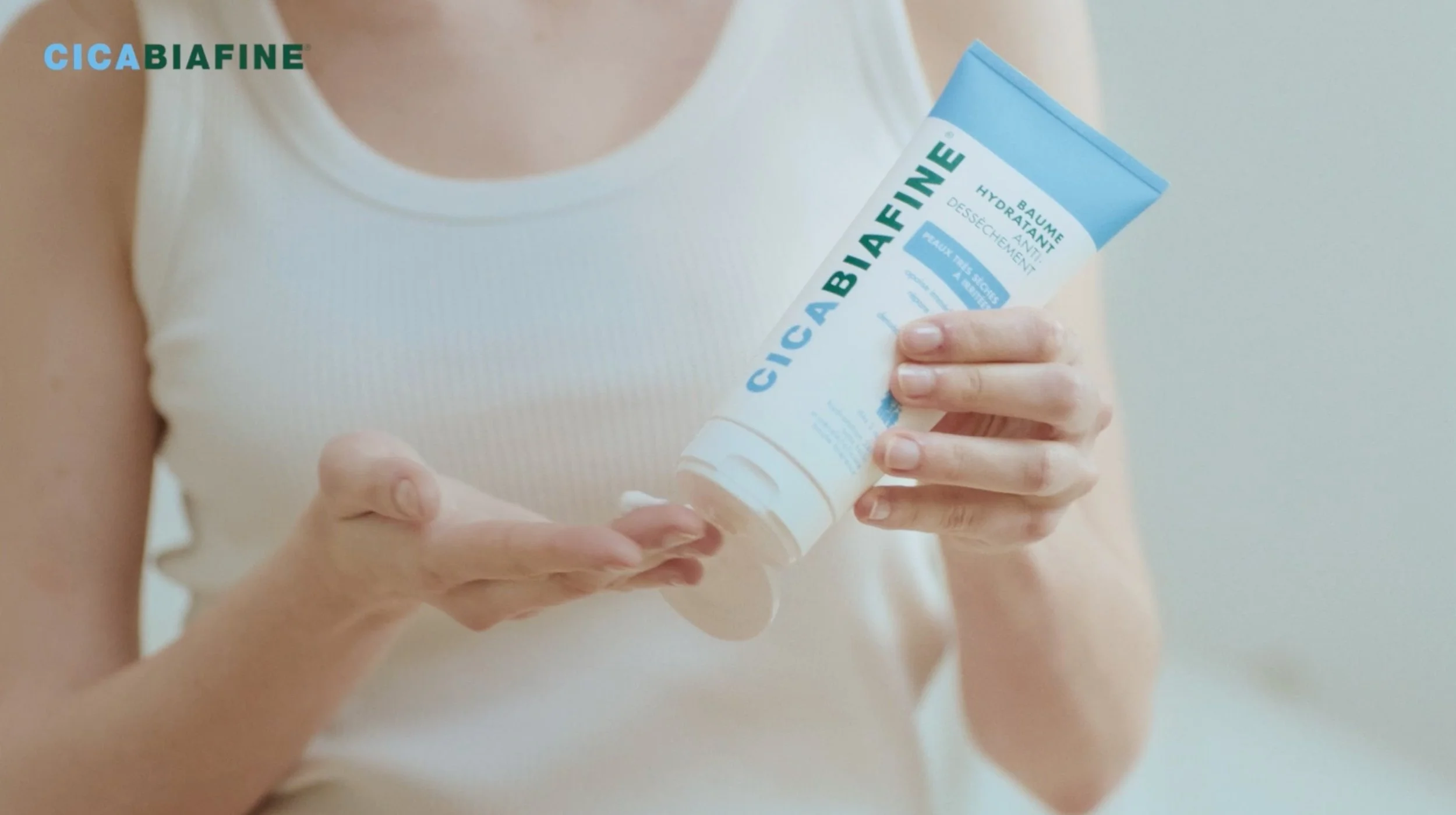 Une personne en débardeur blanc tient un tube de baume hydratant CicaBiafine et en verse une noisette dans la paume de sa main. Le produit est mis en avant sur un fond clair et lumineux
