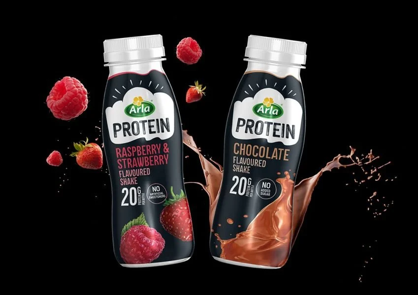 Deux bouteilles de boissons protéinées Arla Protein sont présentées sur un fond noir. L’une est au goût framboise-fraise, l’autre au chocolat. Des fruits et des éclaboussures de chocolat entourent les bouteilles pour illustrer les saveurs.