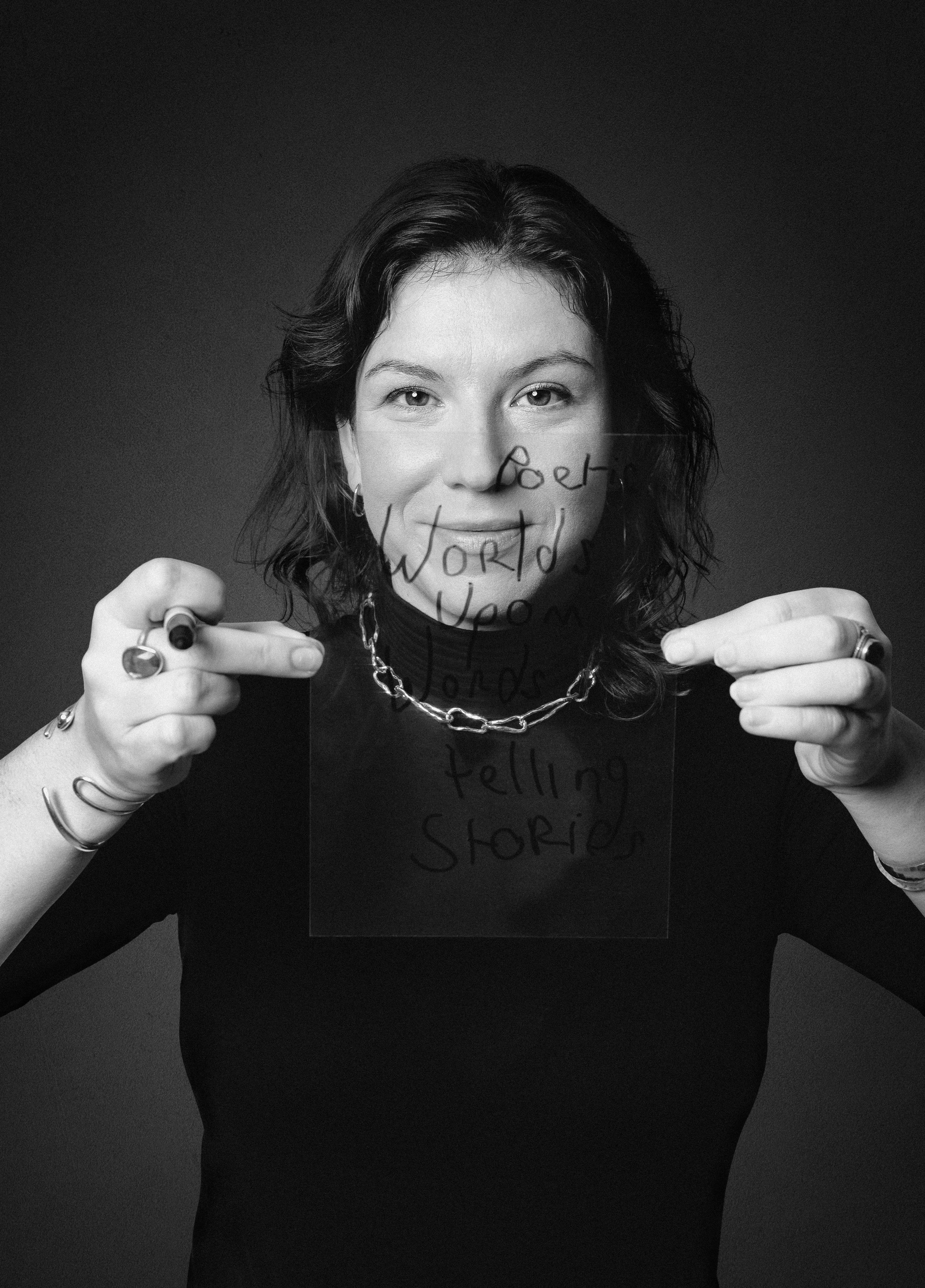 Une femme avec des cheveux foncés et bouclés tient une plaque en verre avec des mots écrits dessus. Elle porte une chaîne métallique autour du cou, plusieurs bagues, et un bracelet. L'image est en noir et blanc.
