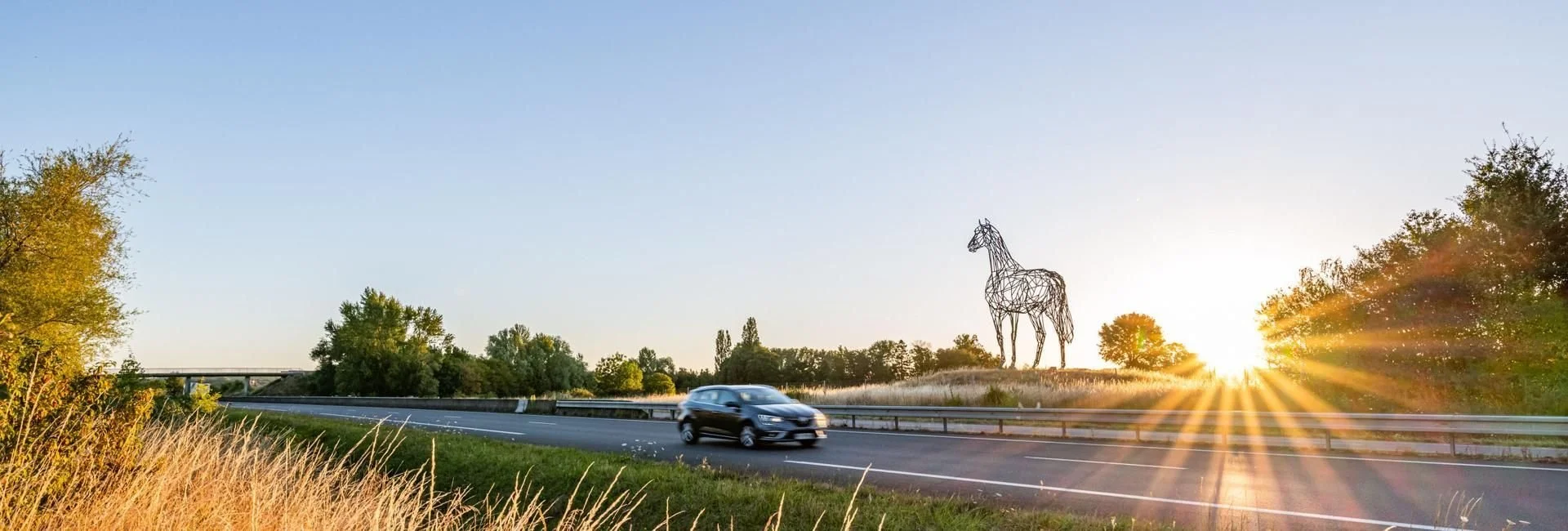 Une voiture noire roule sur une autoroute bordée d’herbes hautes. À droite, une grande sculpture métallique représentant un cheval se dresse dans un champ, avec des arbres en arrière-plan sous un ciel dégagé au coucher du soleil.