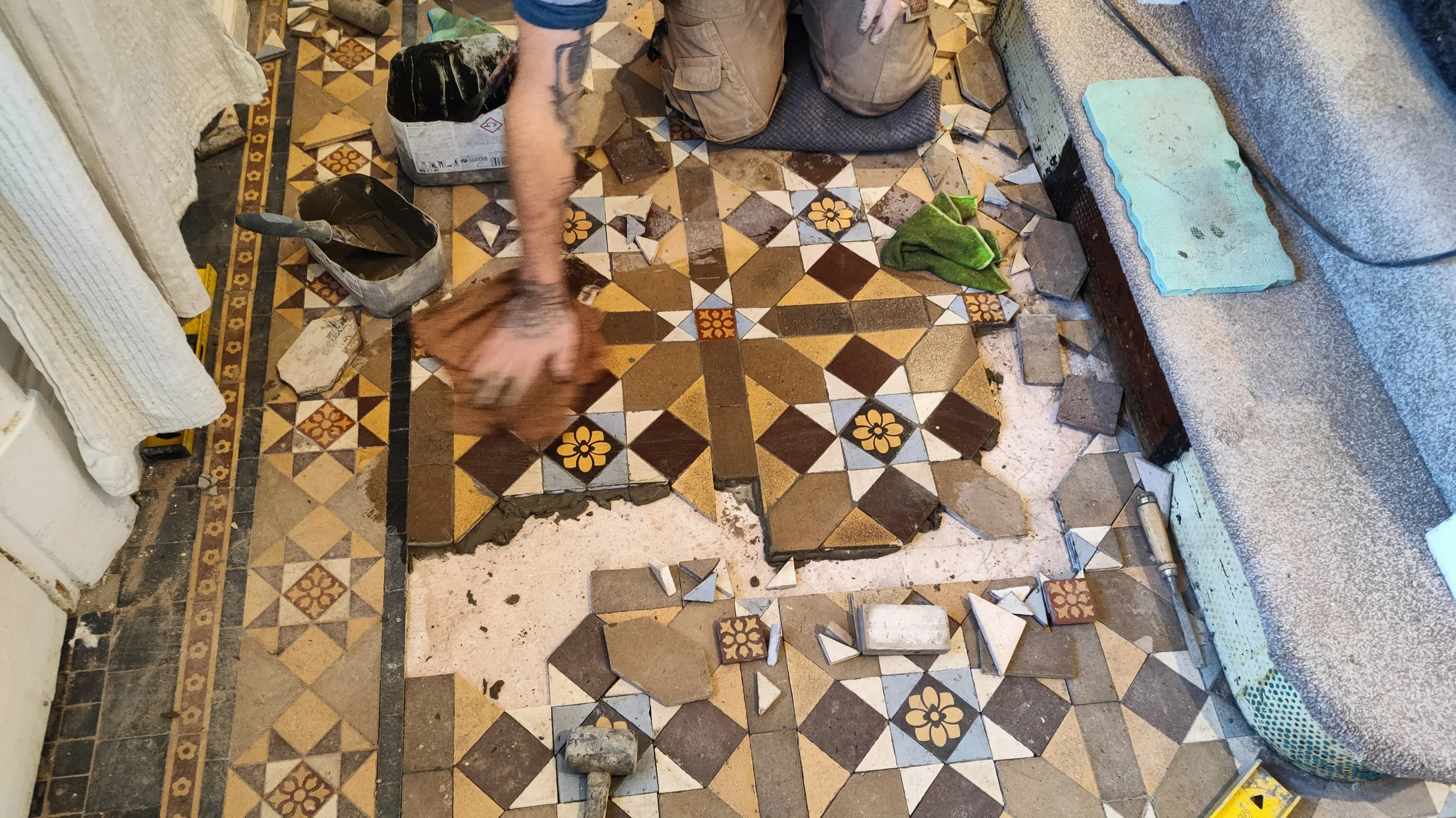A person installing or repairing decorative patterned tile flooring. The person's arm, with tattoos, is visible along with tools and tiles scattered around.