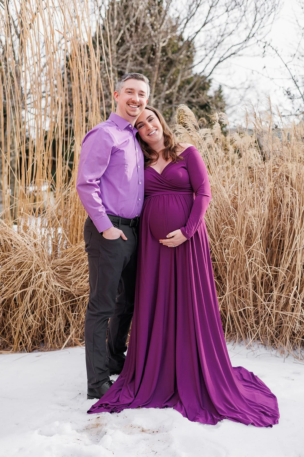 Husband and wife posing together, she is holding her baby bump and resting her head on his shoulder at the Hahn horticulture gardens at Virginia Tech