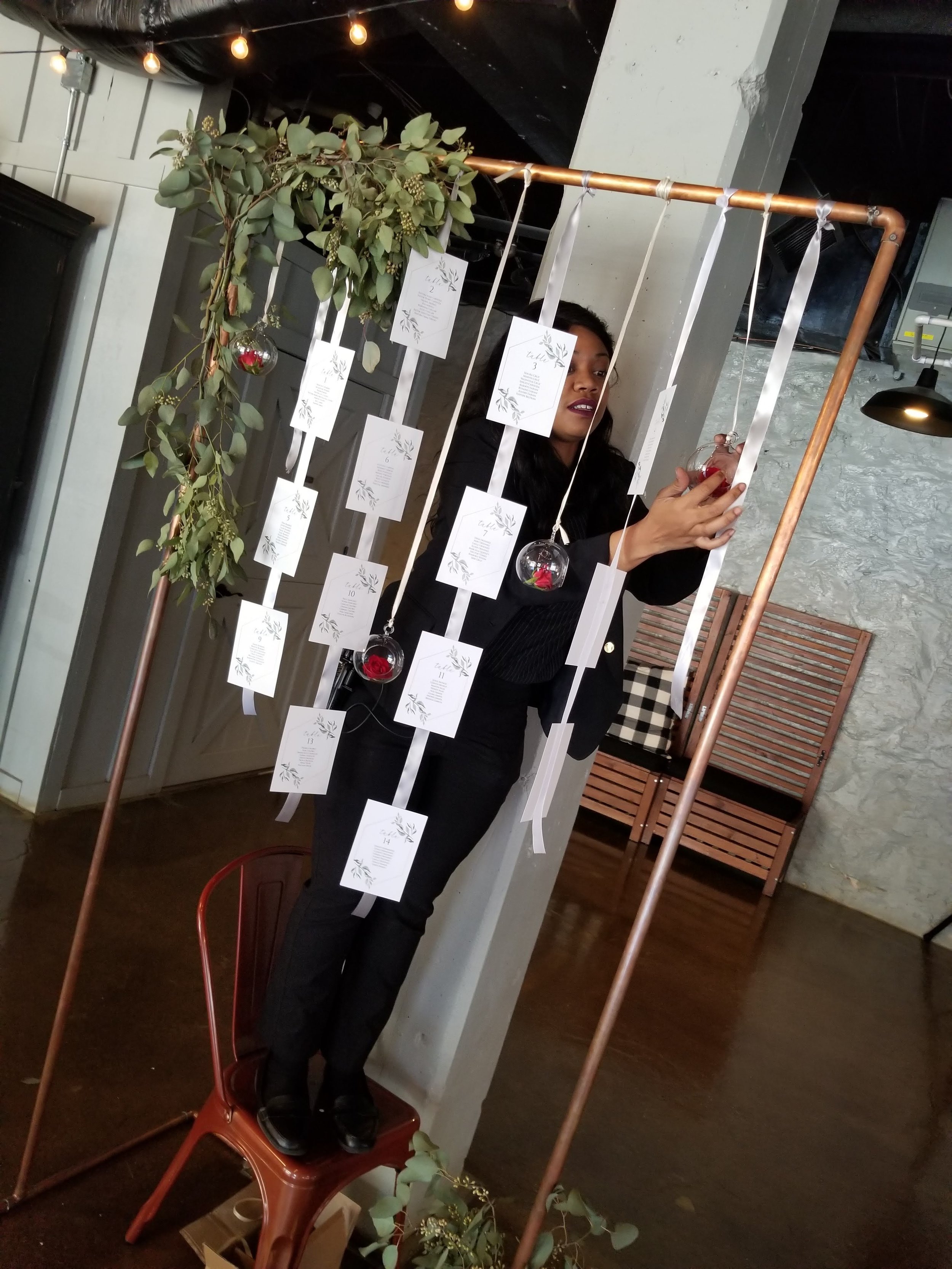 Person standing on a chair setting up a backdrop with hanging white cards and glass ornaments, adorned with greenery, in a decorated indoor space.