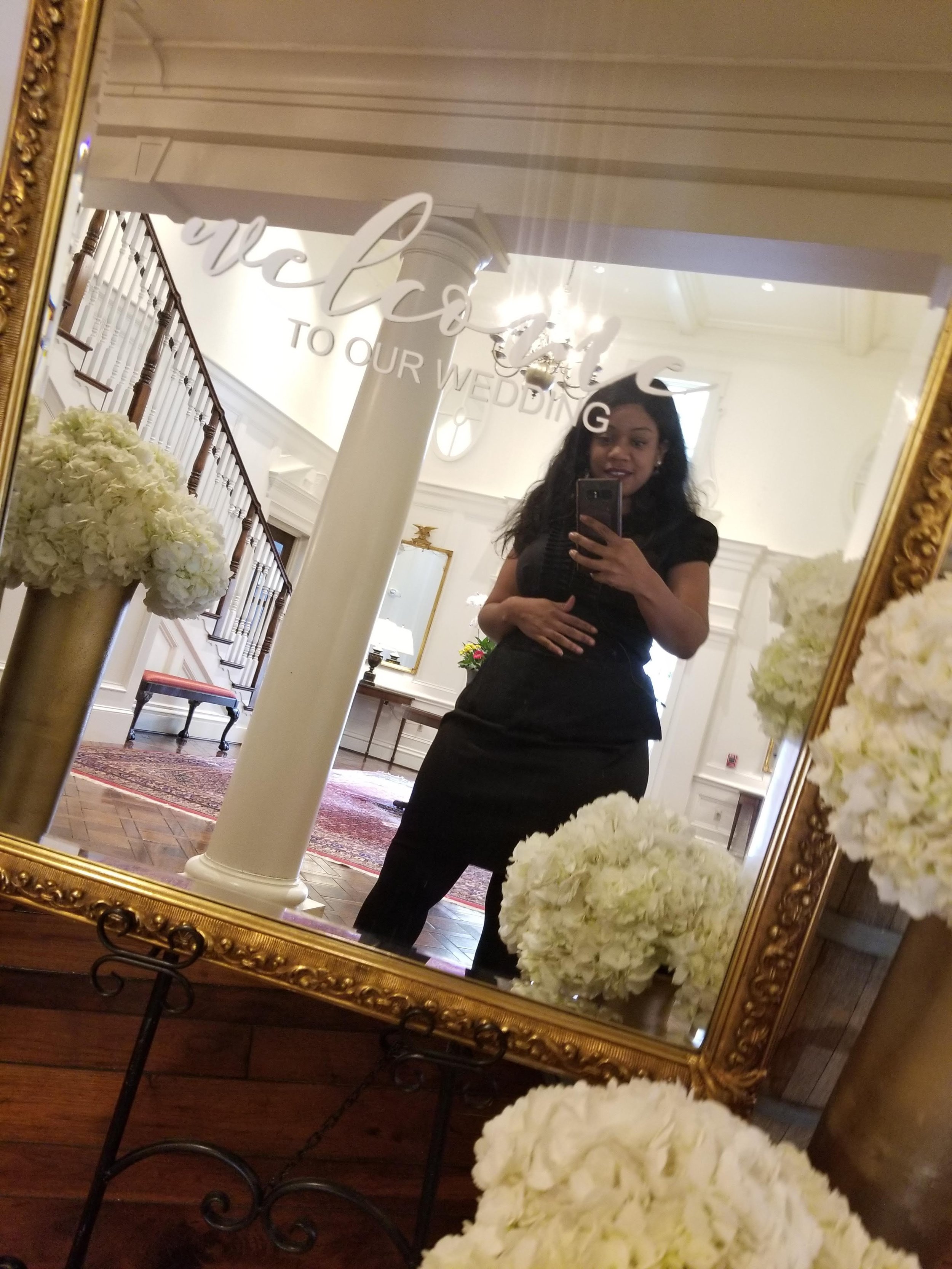 A person taking a mirror selfie at a wedding venue entrance with a gold-framed mirror displaying the text "Welcome to Our Wedding." The room has elegant decor, including white flowers and chandeliers.