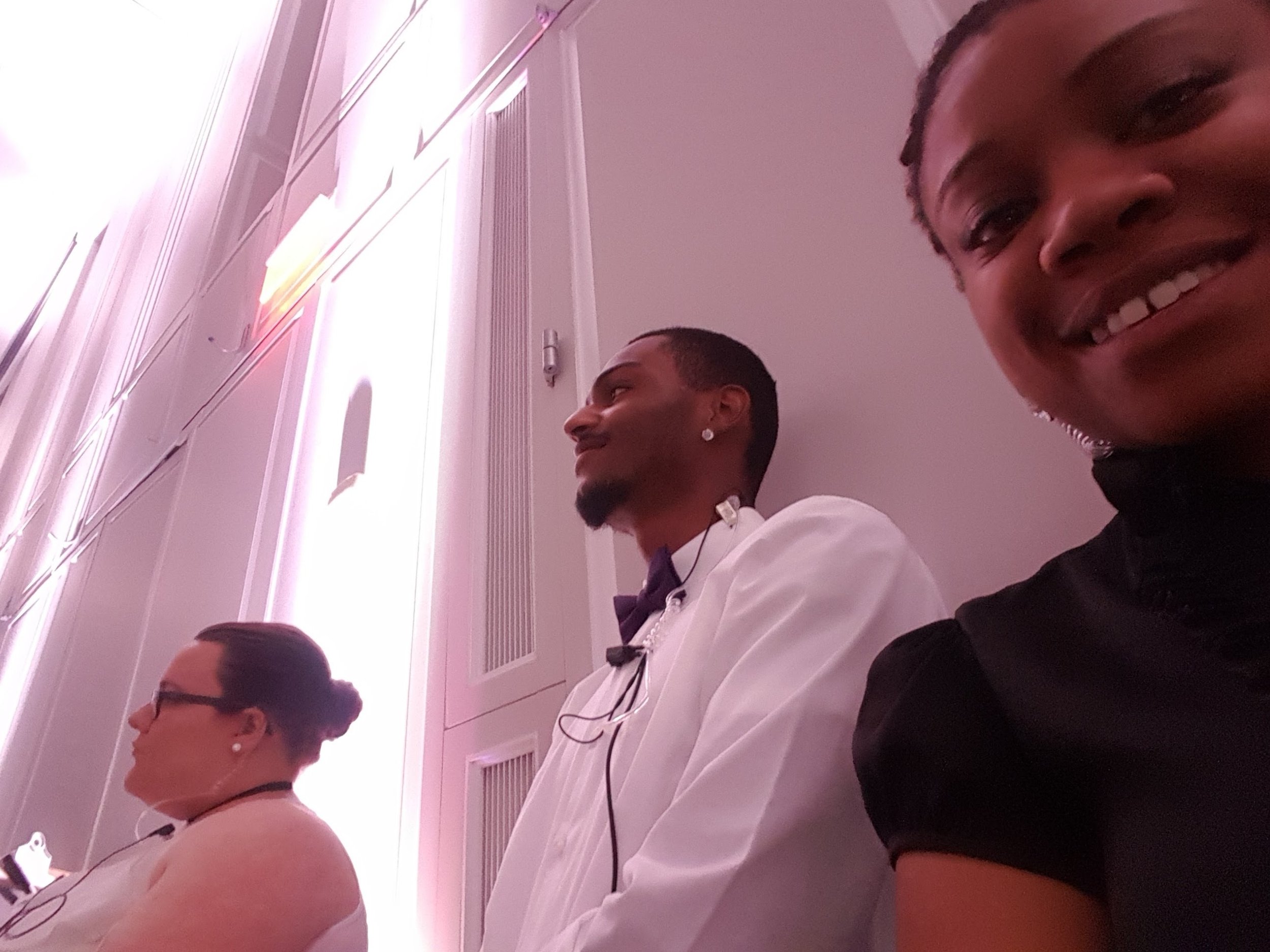 Three people sitting against a wall, with a woman on the right smiling towards the camera. The two on the left are looking away. The setting is indoors with pink lighting.