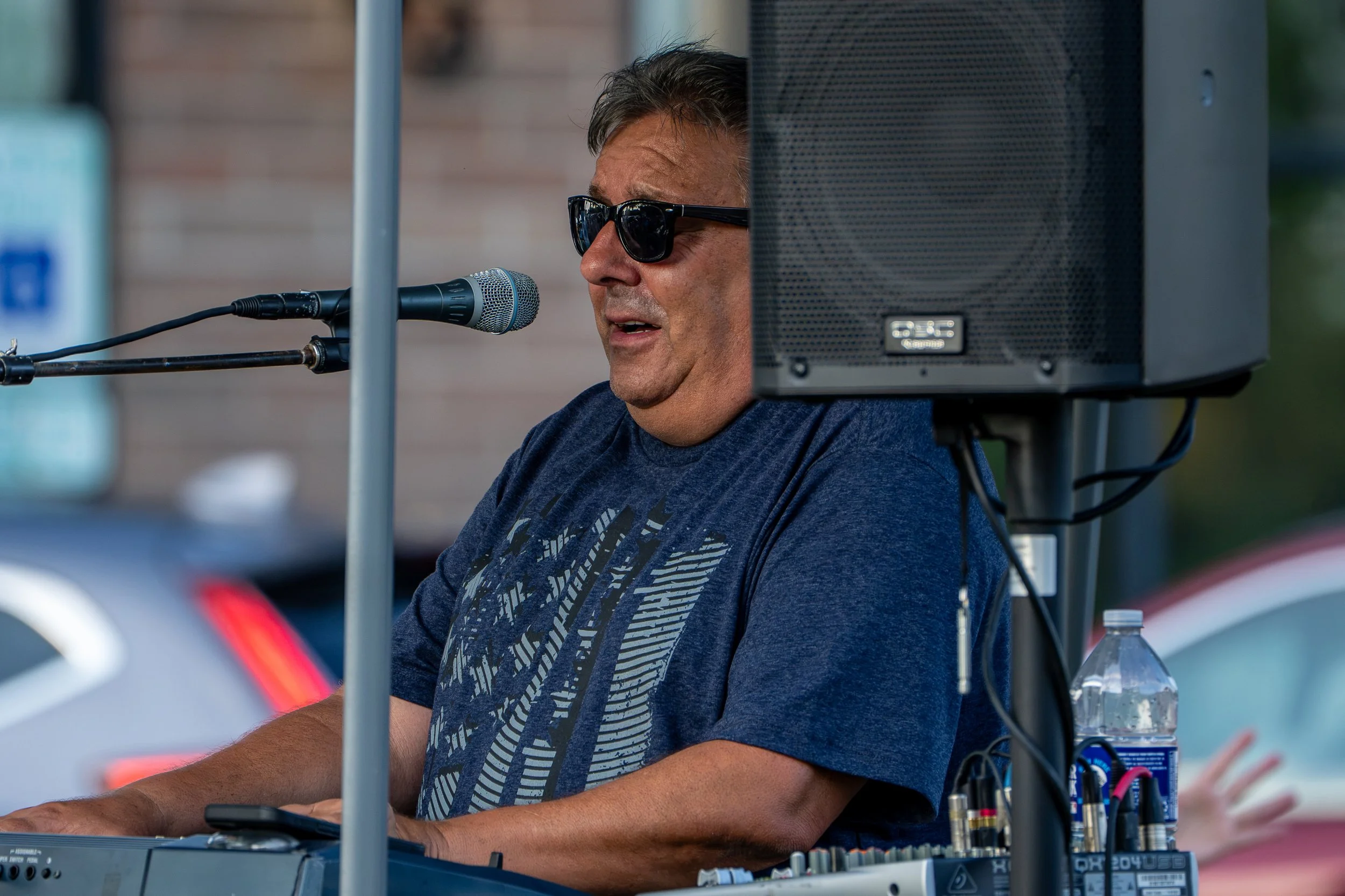A man with short hair, wearing sunglasses and a blue t-shirt, performing music with a keyboard and microphone at an outdoor event.