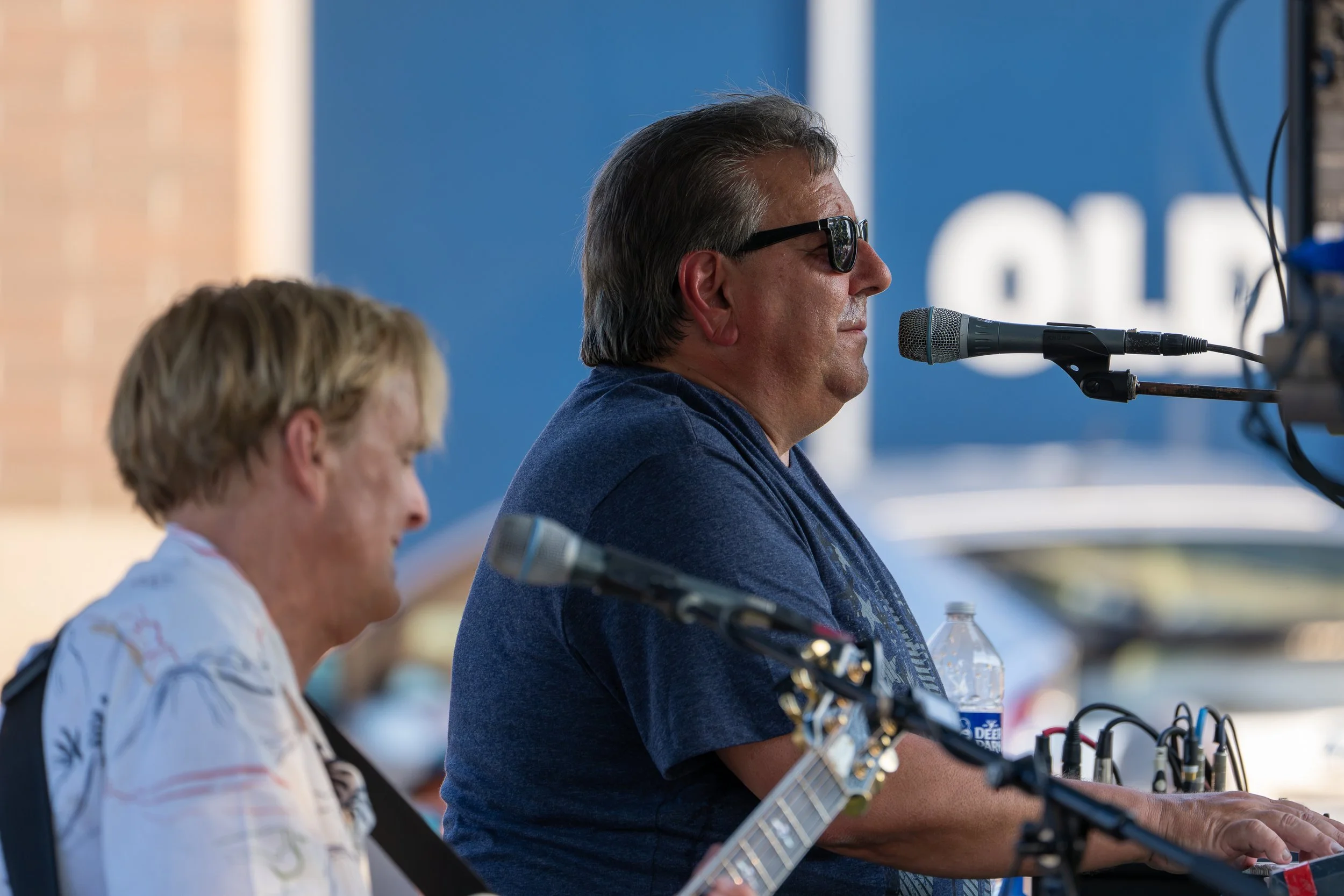 A man wearing sunglasses and a blue t-shirt playing an electric guitar at an outdoor event as part of a band performance.
