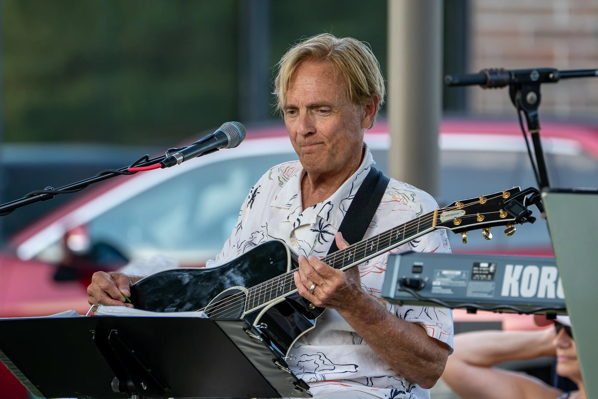A man with blonde hair playing an acoustic guitar at an outdoor event, surrounded by musical equipment.