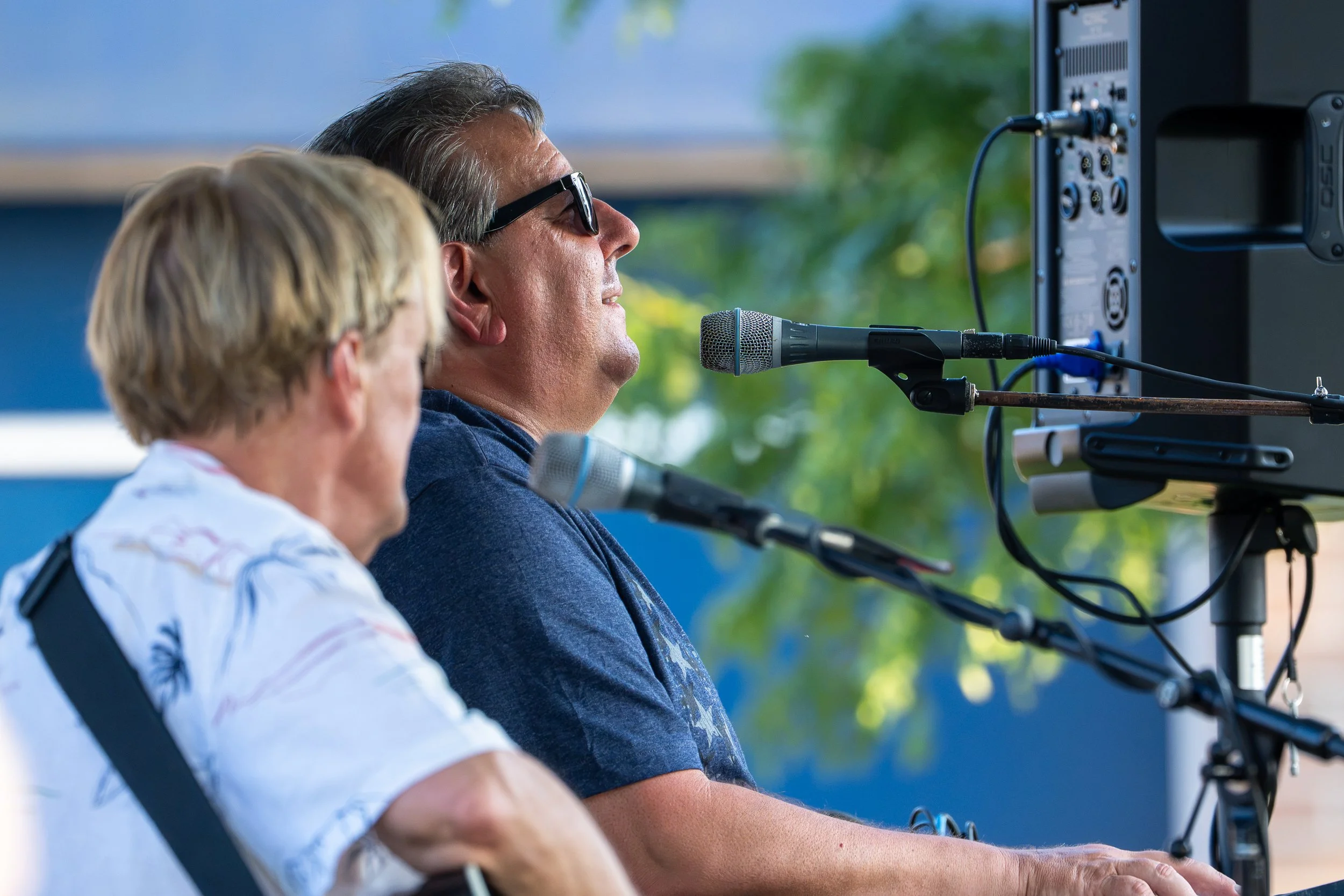 A man and a woman sit side by side outdoors, facing a large monitor with microphones in front of them, appearing to speak or listen. The man, wearing sunglasses and a dark shirt, is looking at the screen, while the woman, with short blonde hair and a