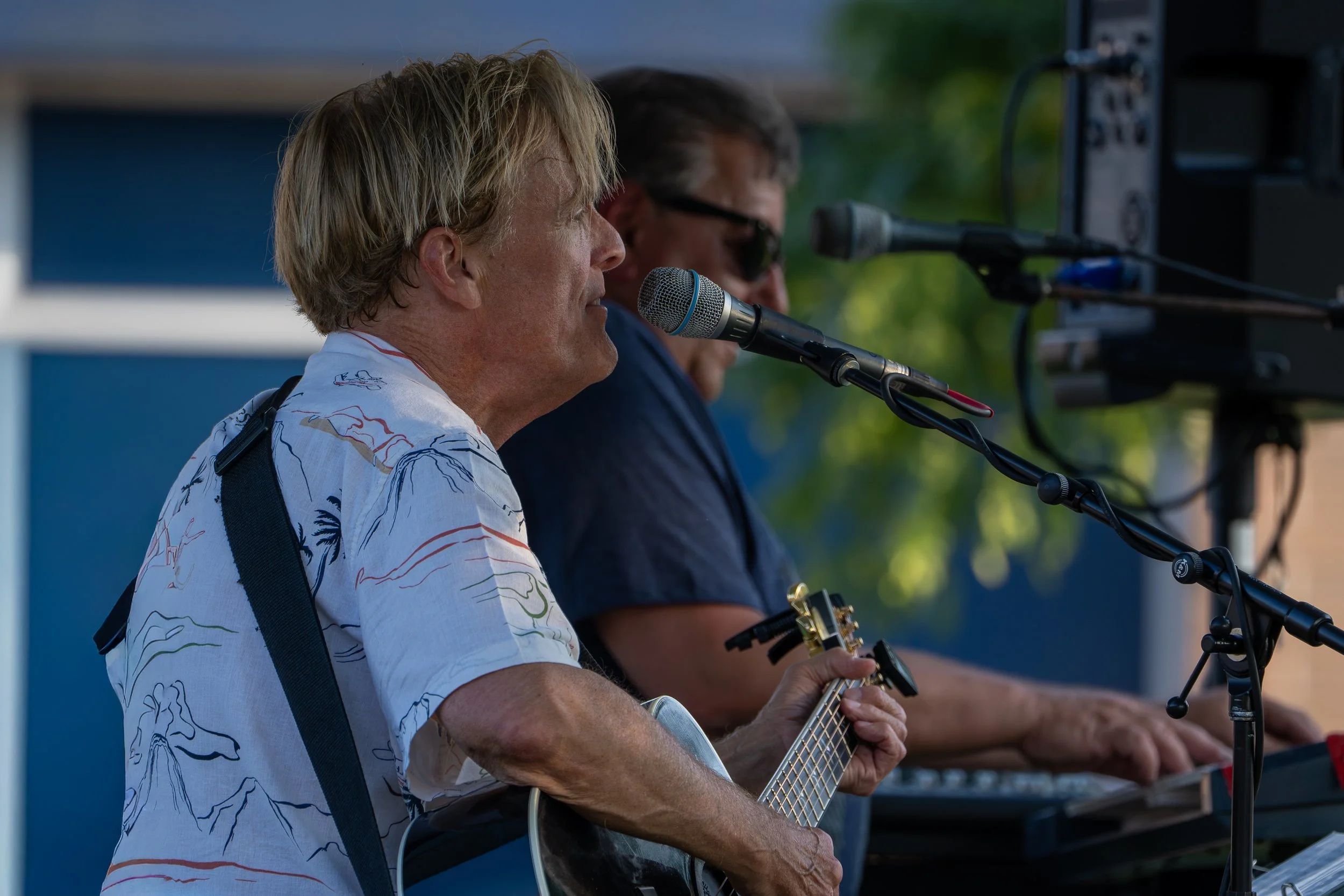 Two men performing music outdoors, one playing a guitar and the other singing or speaking into a microphone, with a background of green trees.