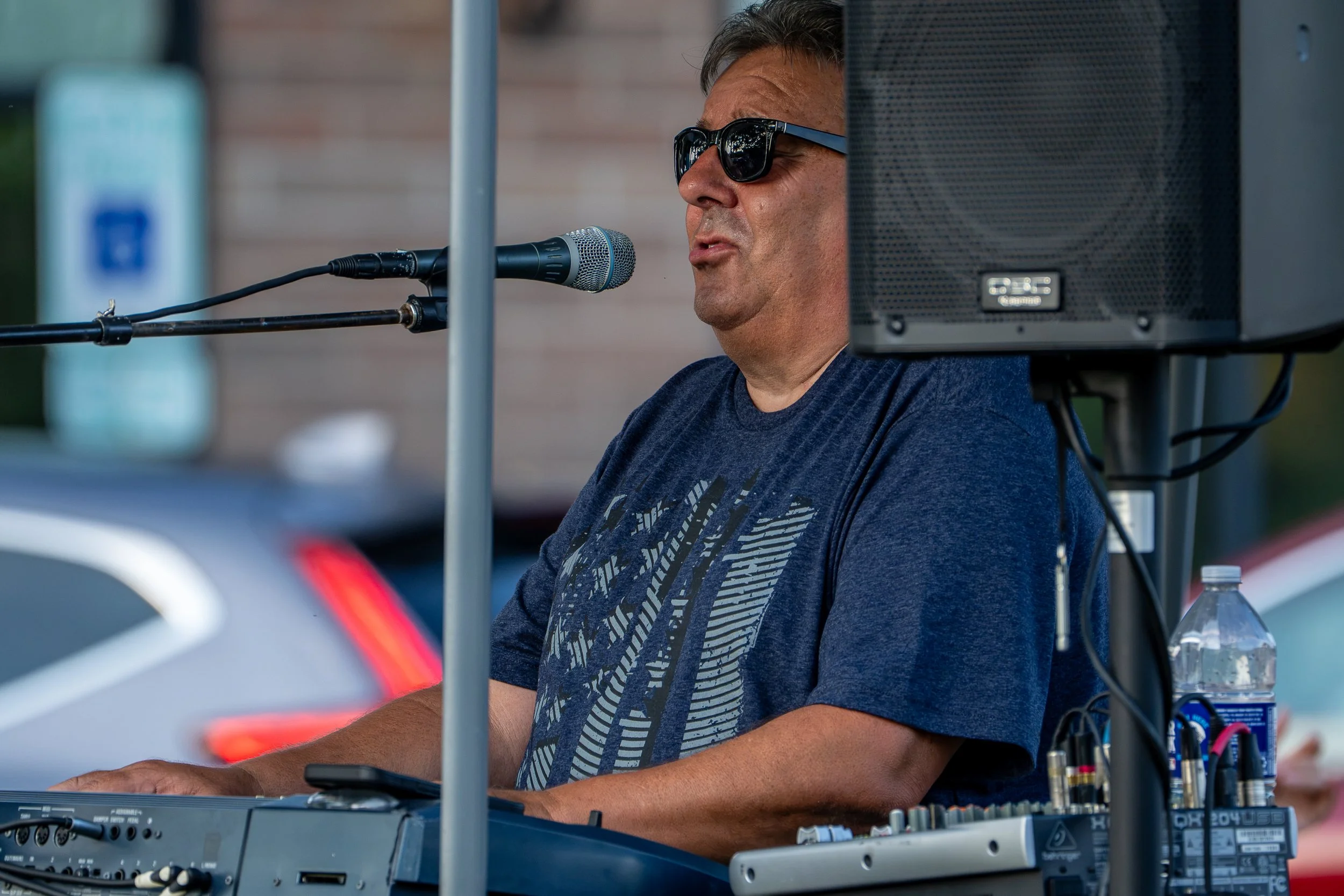 A man wearing sunglasses and a blue t-shirt is speaking into a microphone at an outdoor event. He is positioned behind electronic equipment, possibly a keyboard, with cables connected to it. A plastic water bottle is visible in the lower right corner