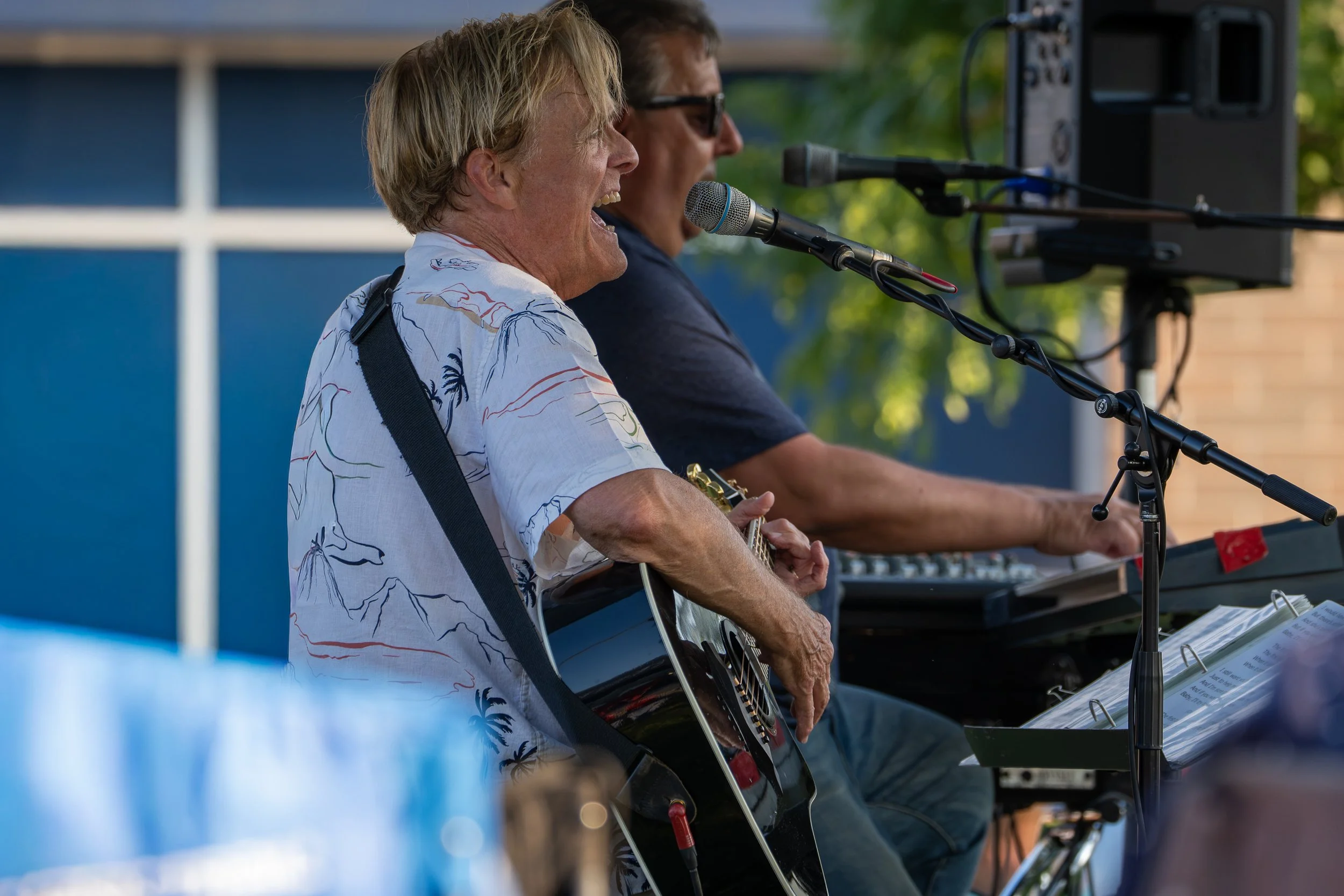 Two men performing music outdoors, one playing guitar and the other singing into a microphone, with audio equipment nearby.