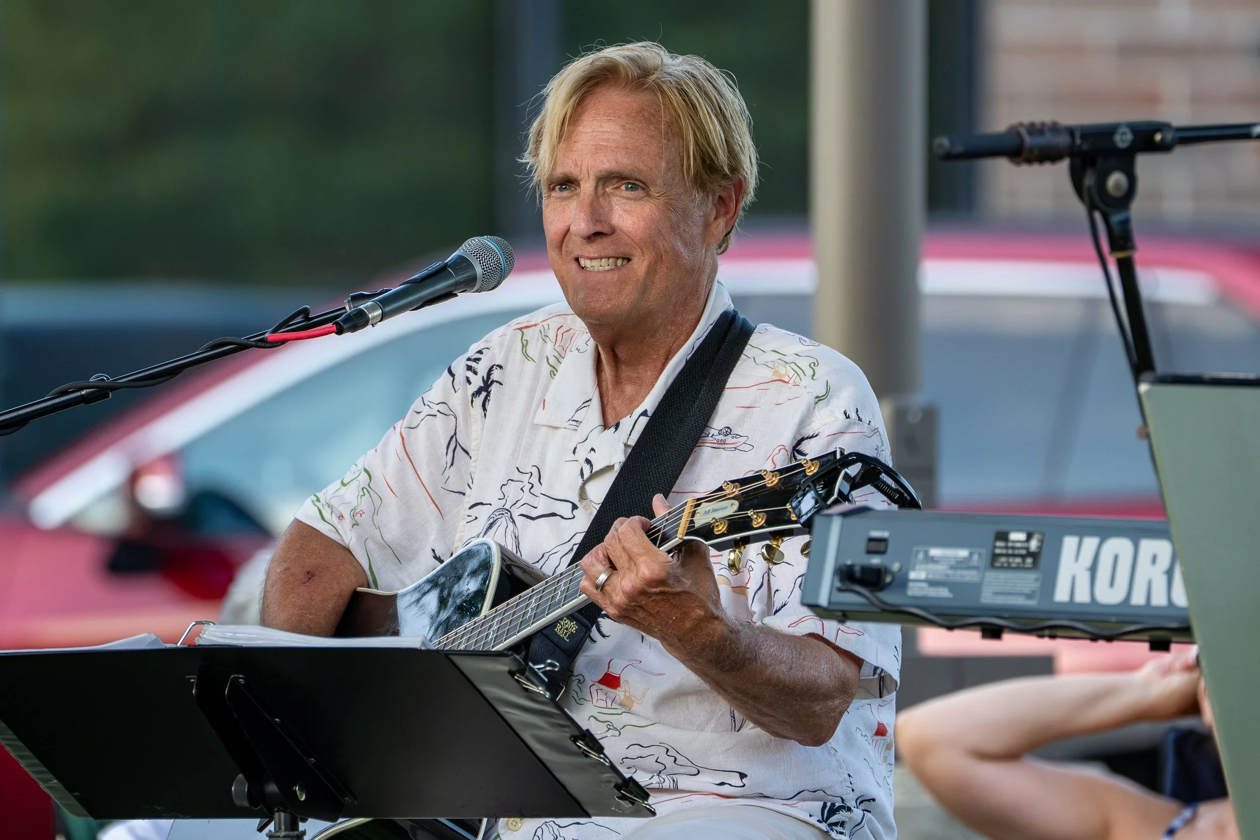 A man with blond hair playing an acoustic guitar and singing into a microphone during an outdoor performance, with musical equipment around him.