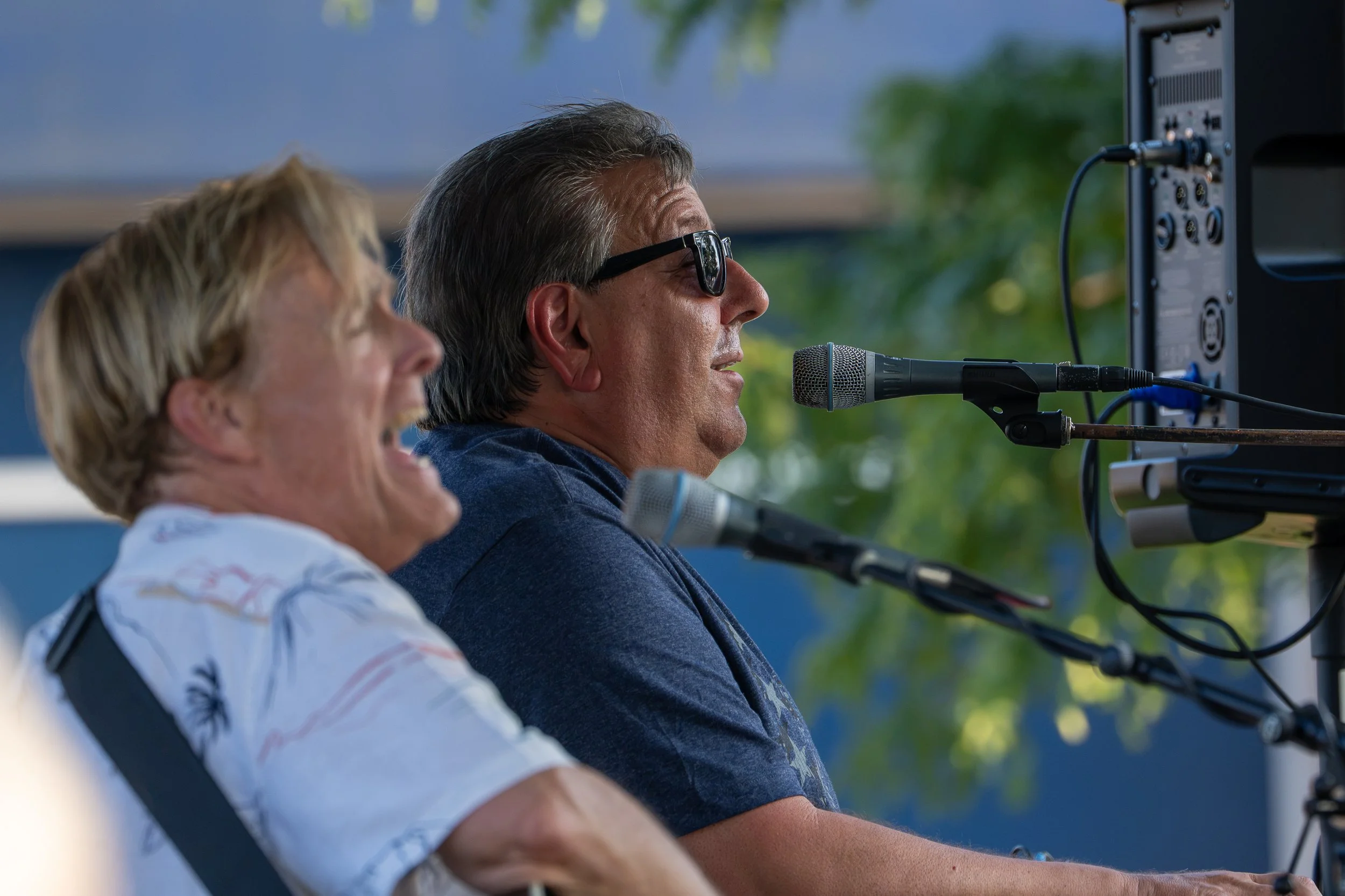 A man wearing glasses speaking into a microphone at an outdoor event, with a woman next to him also speaking into a microphone.