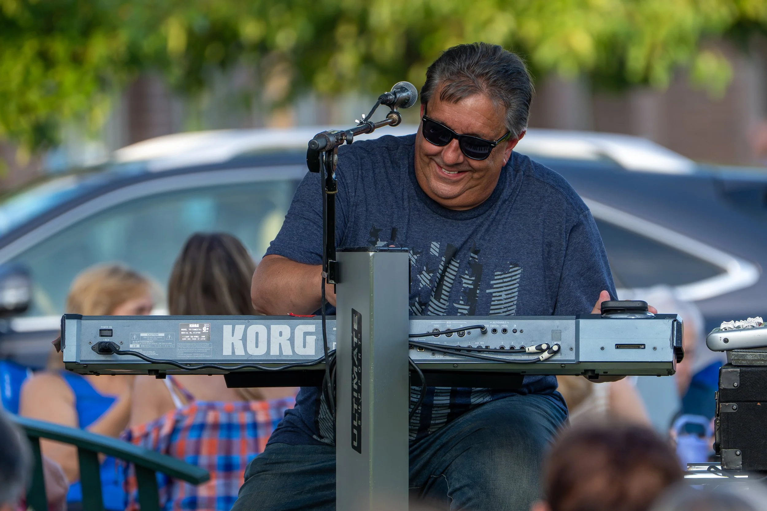 A man wearing sunglasses and a blue T-shirt playing a keyboard on a stand outdoors during the daytime, with children and cars in the background.