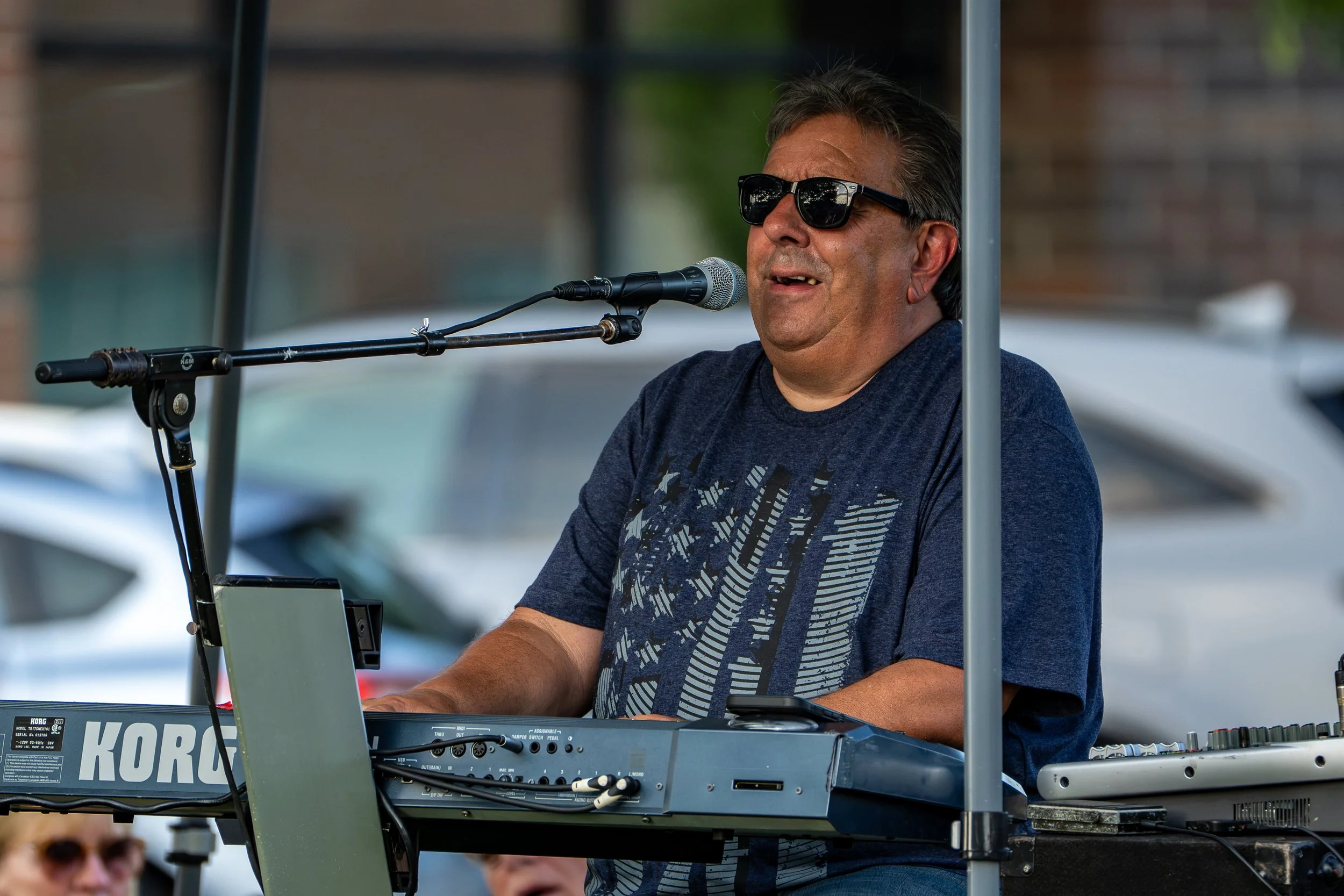 A man wearing sunglasses is playing a keyboard and singing into a microphone at an outdoor event.