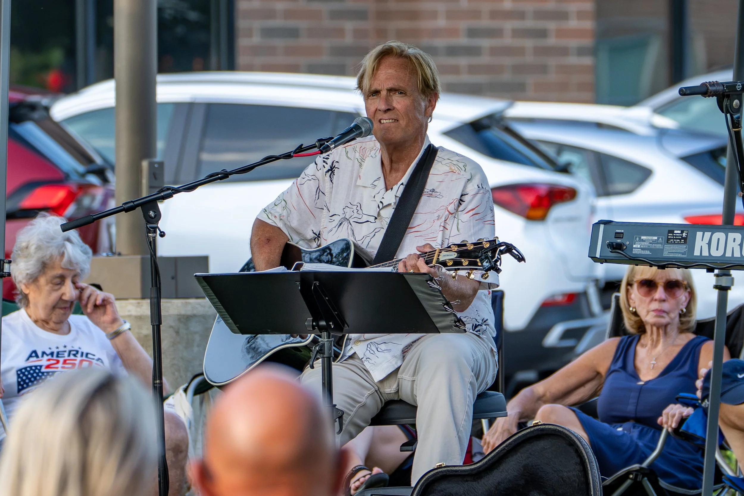 A man playing guitar and singing into a microphone outdoors during a gathering, with several people seated around him, some wearing patriotic-themed clothing.