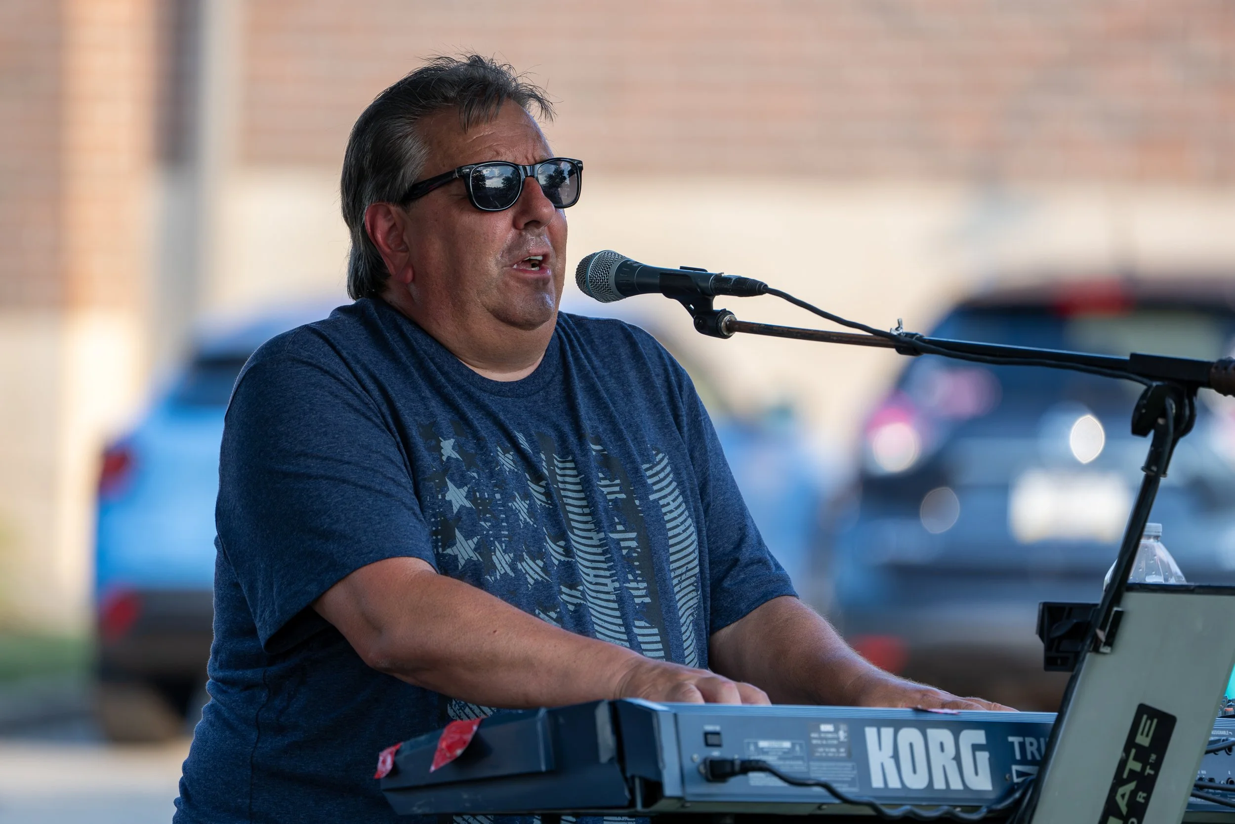 A man wearing glasses playing a keyboard and singing into a microphone outdoors.