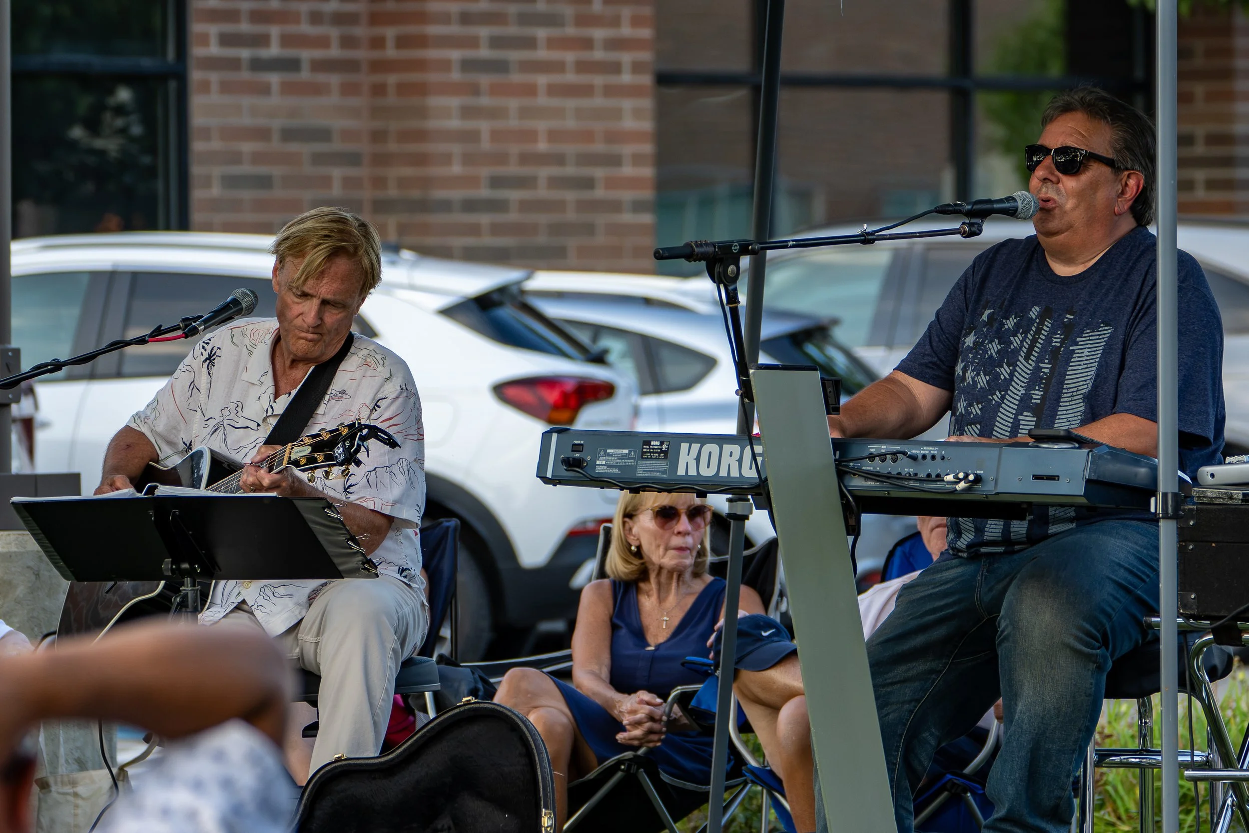 Musicians performing at an outdoor event, including a man playing guitar and a man singing into a microphone with a keyboard in front of him. Audience members are seated, including elderly women, with cars and a brick building in the background.