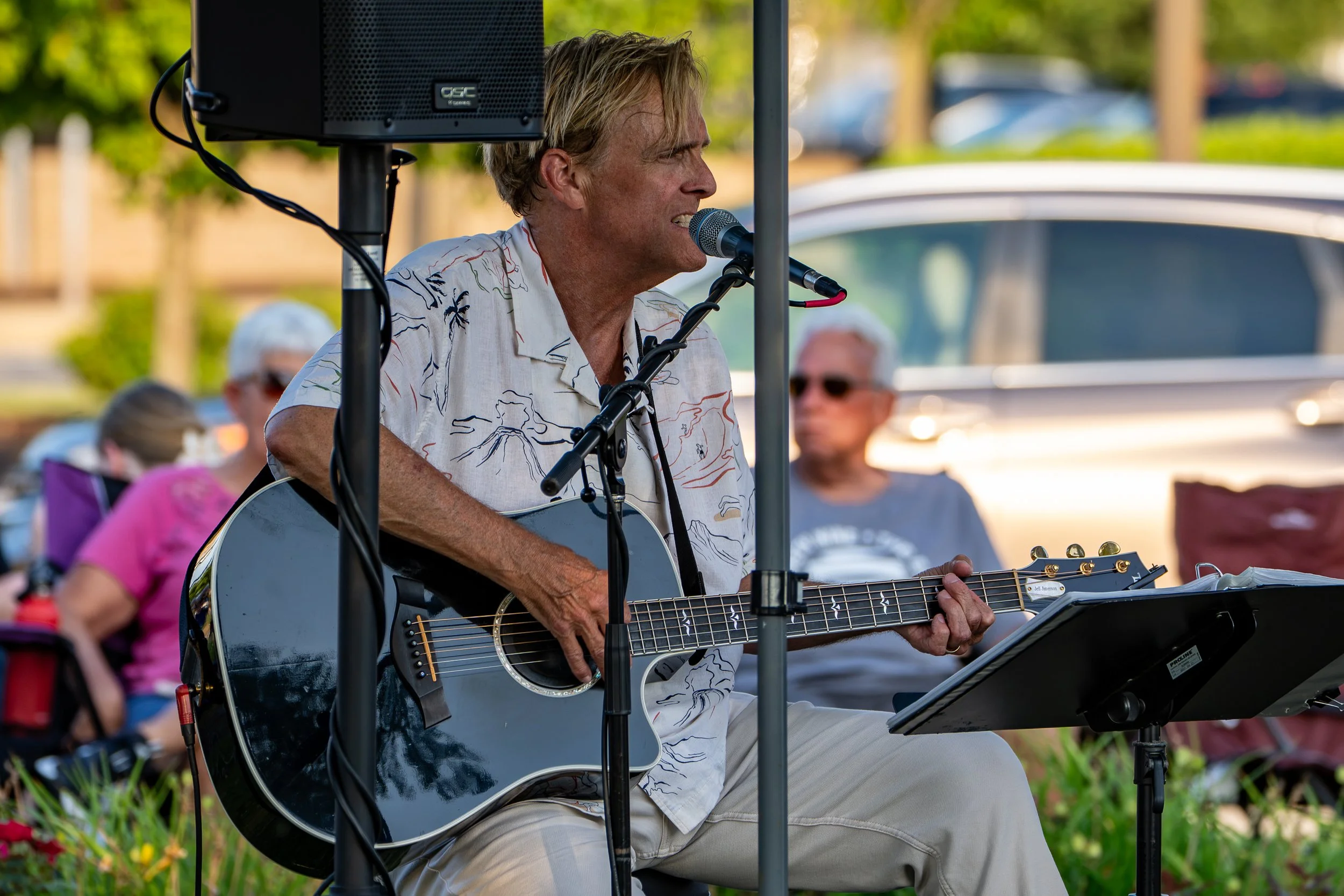 A man with light brown hair, wearing a Hawaiian shirt, is singing into a microphone while playing an acoustic guitar at an outdoor event. There are blurred spectators in the background.