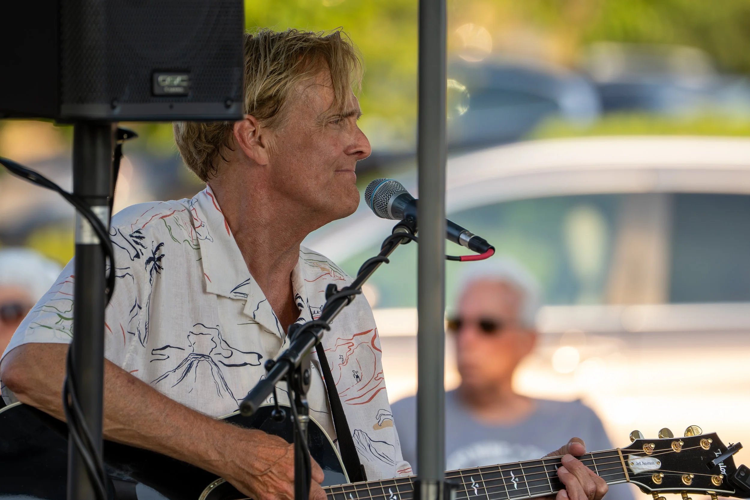 A man with blond hair playing an acoustic guitar and singing into a microphone at an outdoor event, with blurred background and audience members visible.