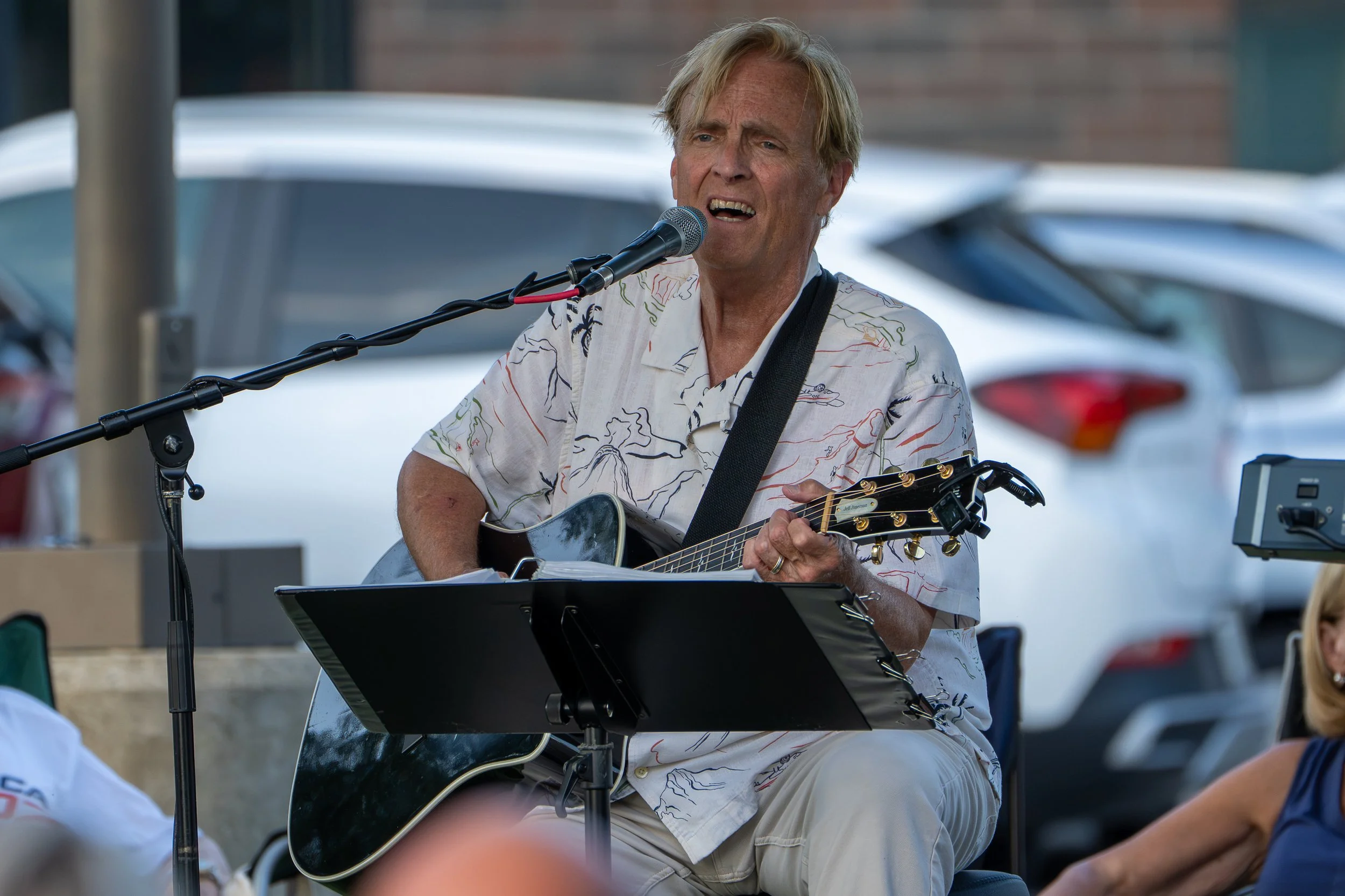 A middle-aged man with blond hair singing into a microphone while playing an acoustic guitar at an outdoor event. He is wearing a white shirt with colorful lines and appears to be emotionally engaged in his performance.