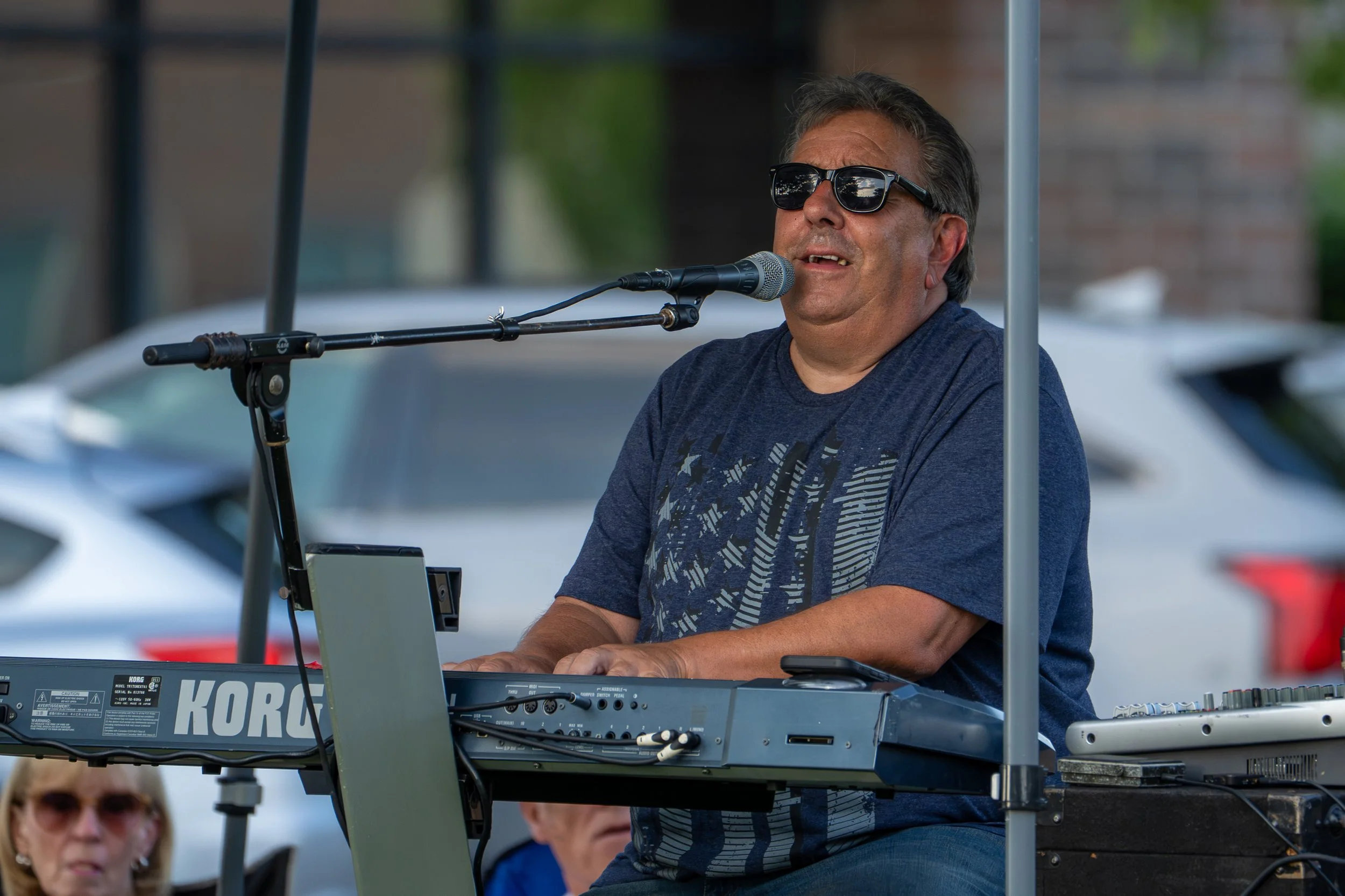 A man wearing sunglasses and a navy blue T-shirt singing into a microphone while playing a keyboard on an outdoor stage.