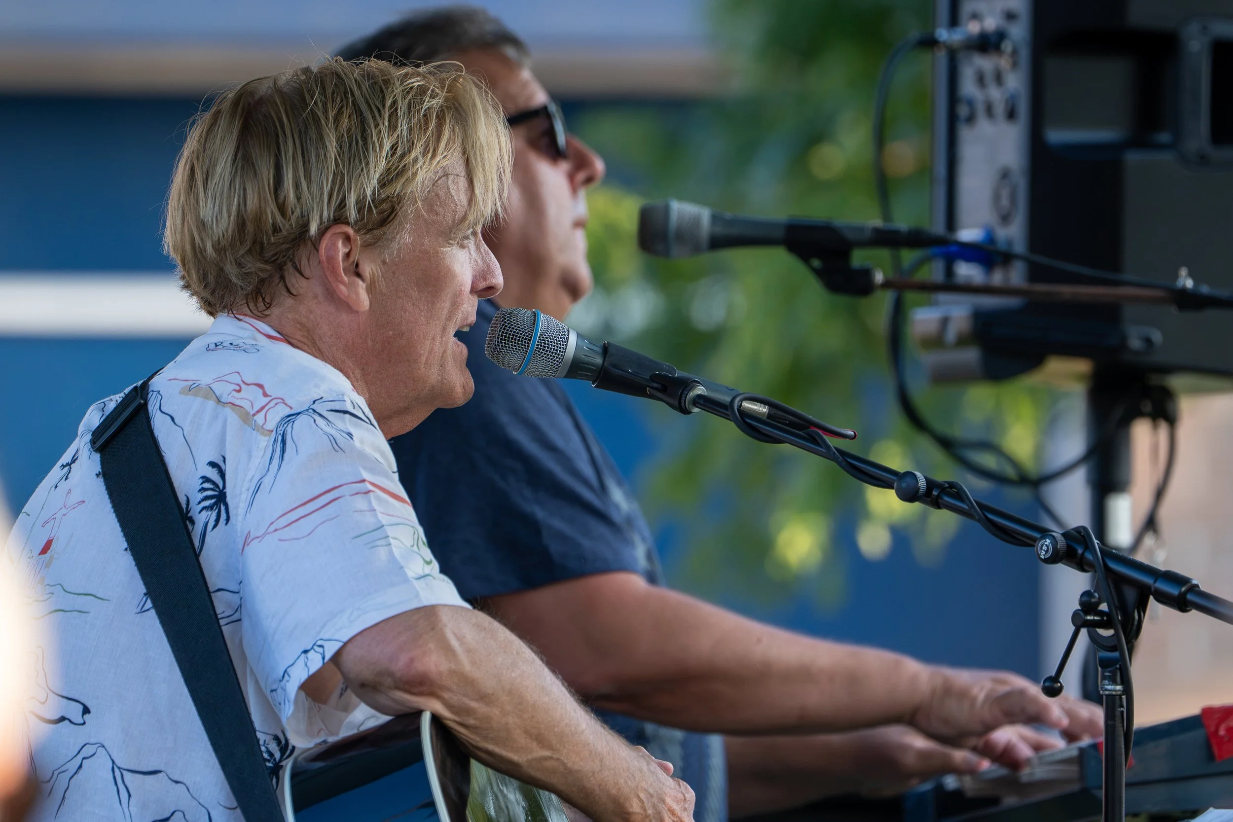 Two people are playing the keyboard outdoors, with one wearing sunglasses and a dark shirt, and the other in a white shirt with colorful prints. They are singing into microphones while sitting at a musical setup.
