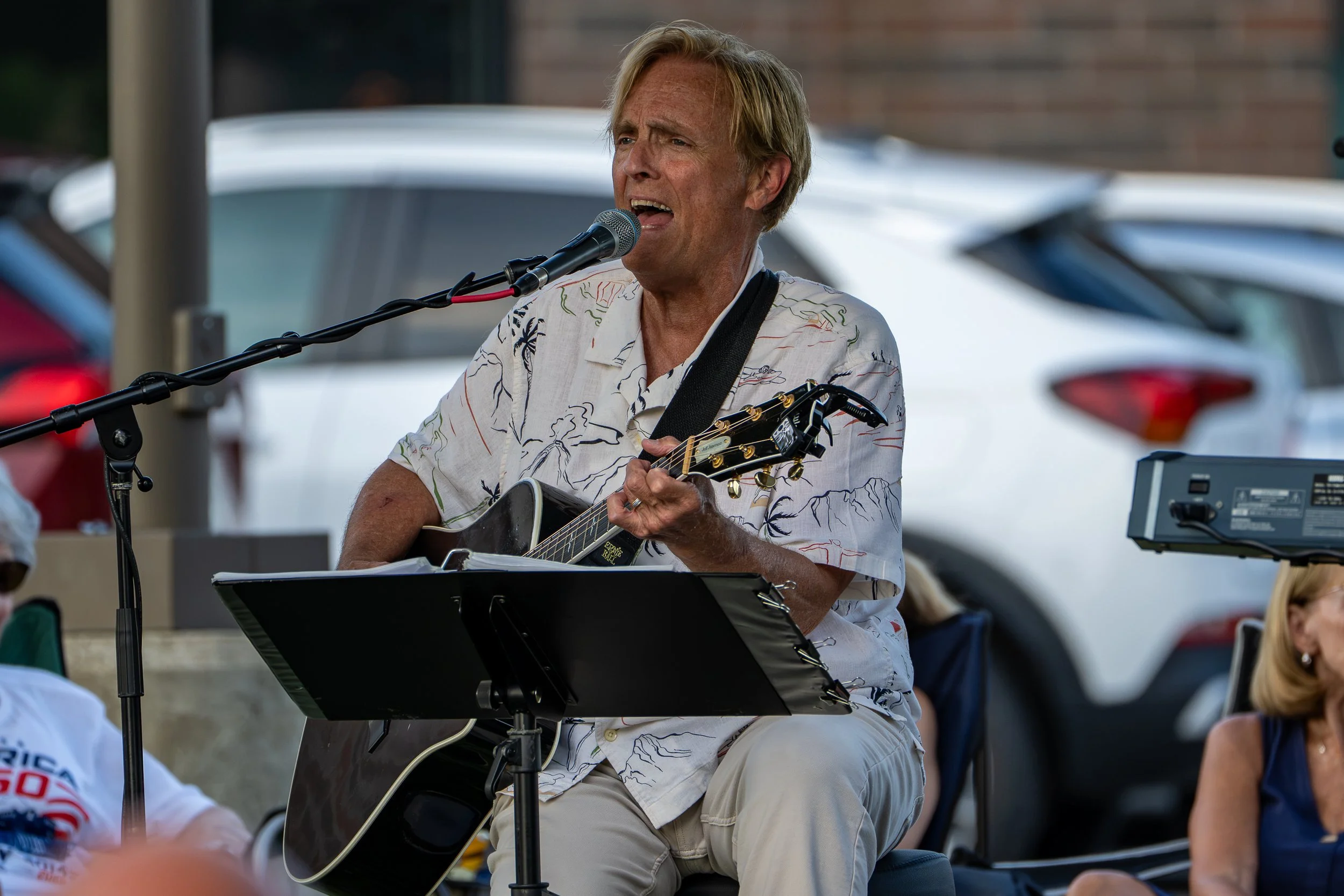 A man with blonde hair and a floral shirt singing into a microphone while playing an acoustic guitar during an outdoor event.