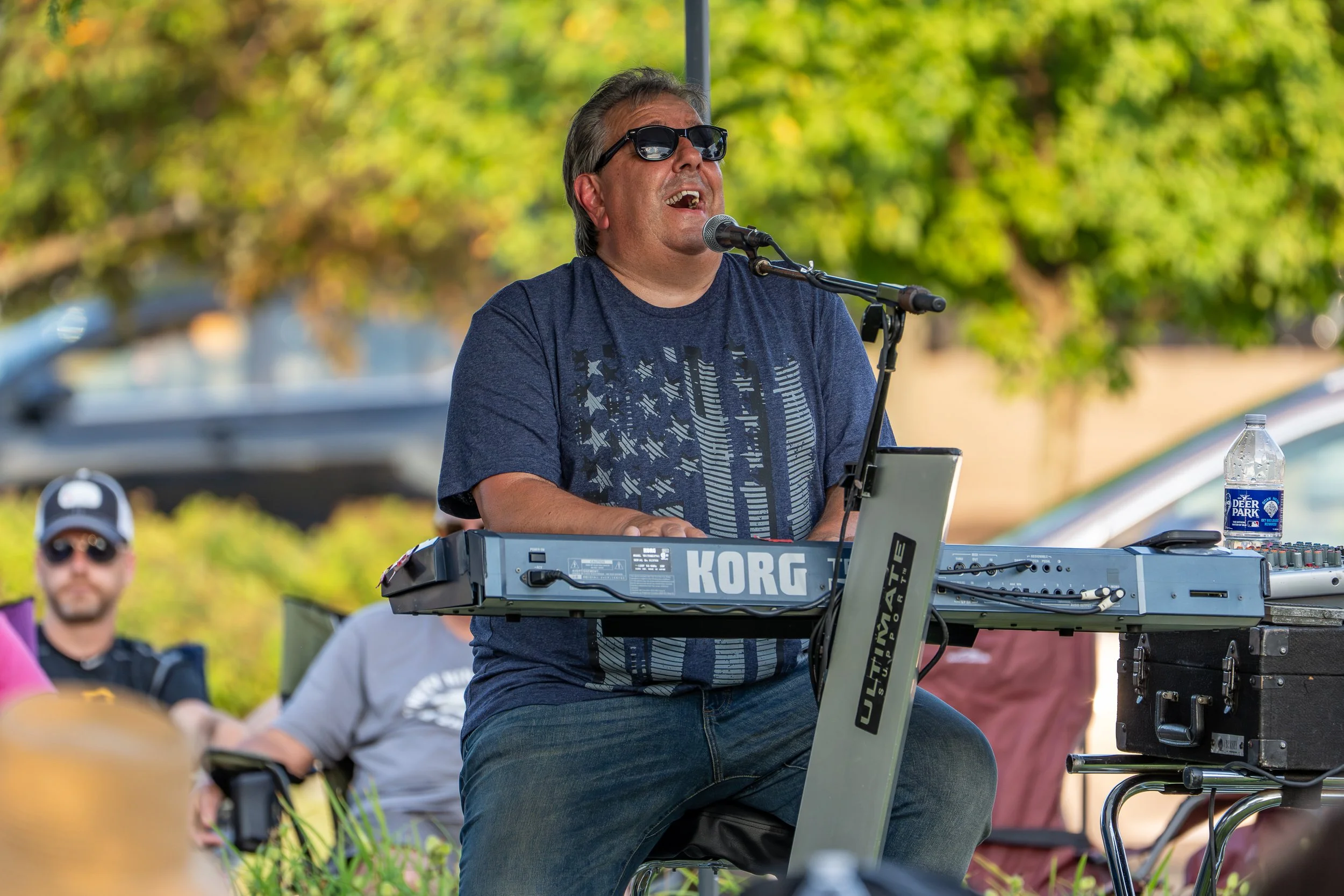 A man wearing sunglasses singing into a microphone while playing a Korg keyboard outdoors, with a green leafy background.